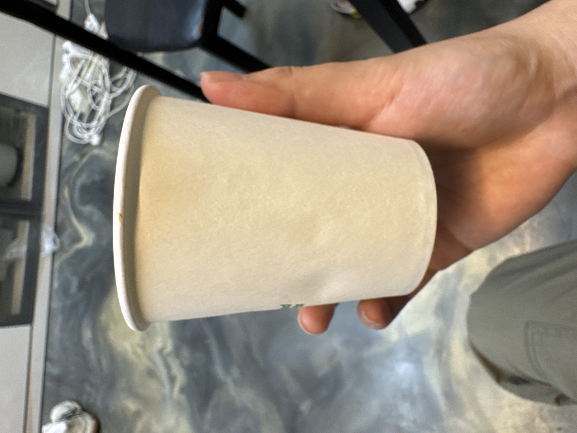 A person is holding a white paper cup. The location is Hwaseong-si, South Korea. The person is wearing khaki-colored pants. In the background, there is a black chair and a tangled white cord on a gray surface.