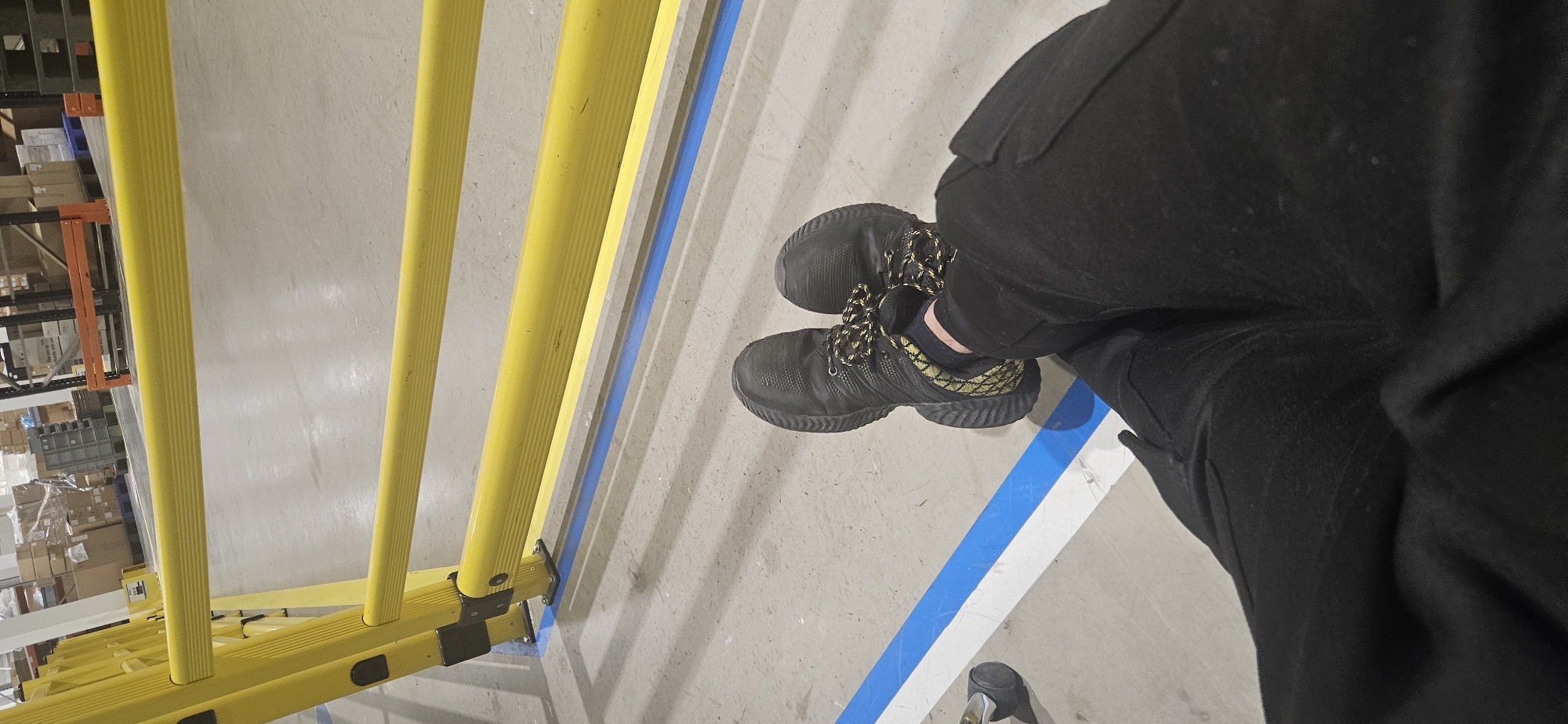 The image depicts a person standing on a concrete floor, next to a yellow ladder. The person is wearing black pants and dark-colored shoes with yellow and black laces. A blue and white stripe is painted on the floor. The location appears to be a warehouse or industrial space, suggested by the presence of shelves and stored items in the background. The location is not in the city of Plovdiv, Bulgaria.