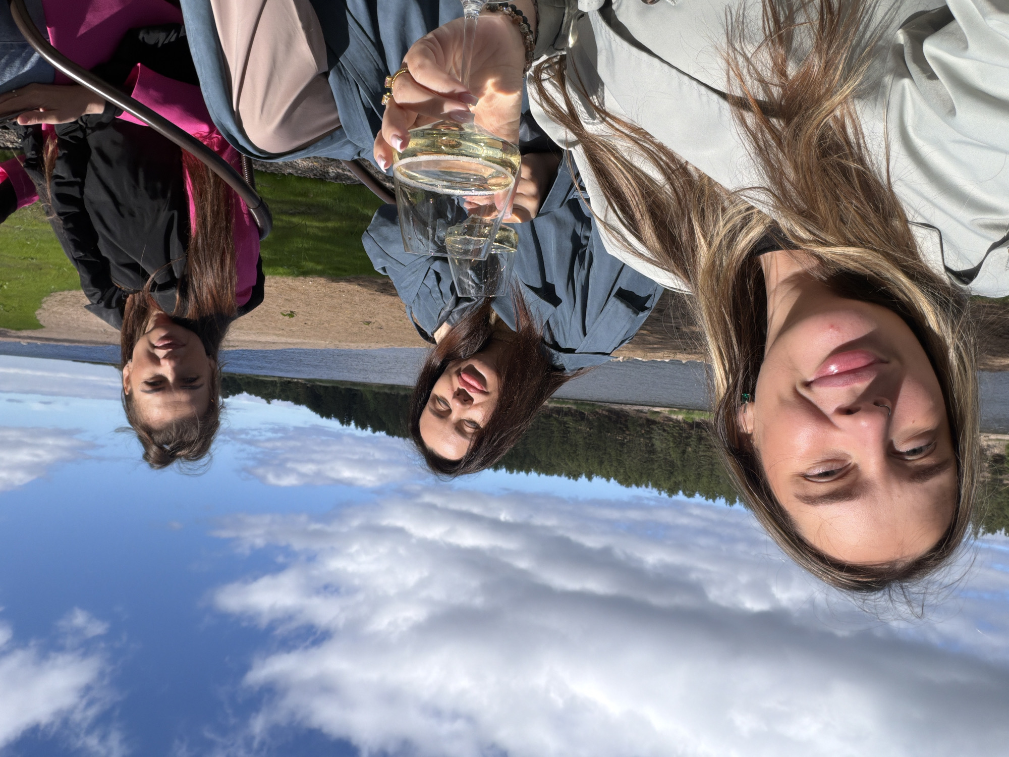 Three women are pictured outdoors in Solov'evo, Russia, near a body of water. The woman in the foreground on the left is facing the camera, smiling, and her shoulders and upper chest are visible. She has long, blonde-brown hair and is wearing a light-colored trench coat over a black shirt. To her right, a woman in the middle is leaning back with her eyes closed, holding a wine glass filled with white wine. She is wearing a blue trench coat. Another woman is seated to the right, holding a wine glass and looking towards the camera. She has long brown hair and is wearing a black jacket over a pink garment. The background consists of a sandy shore, green grass, and a dense forest of pine trees against a cloudy blue sky.