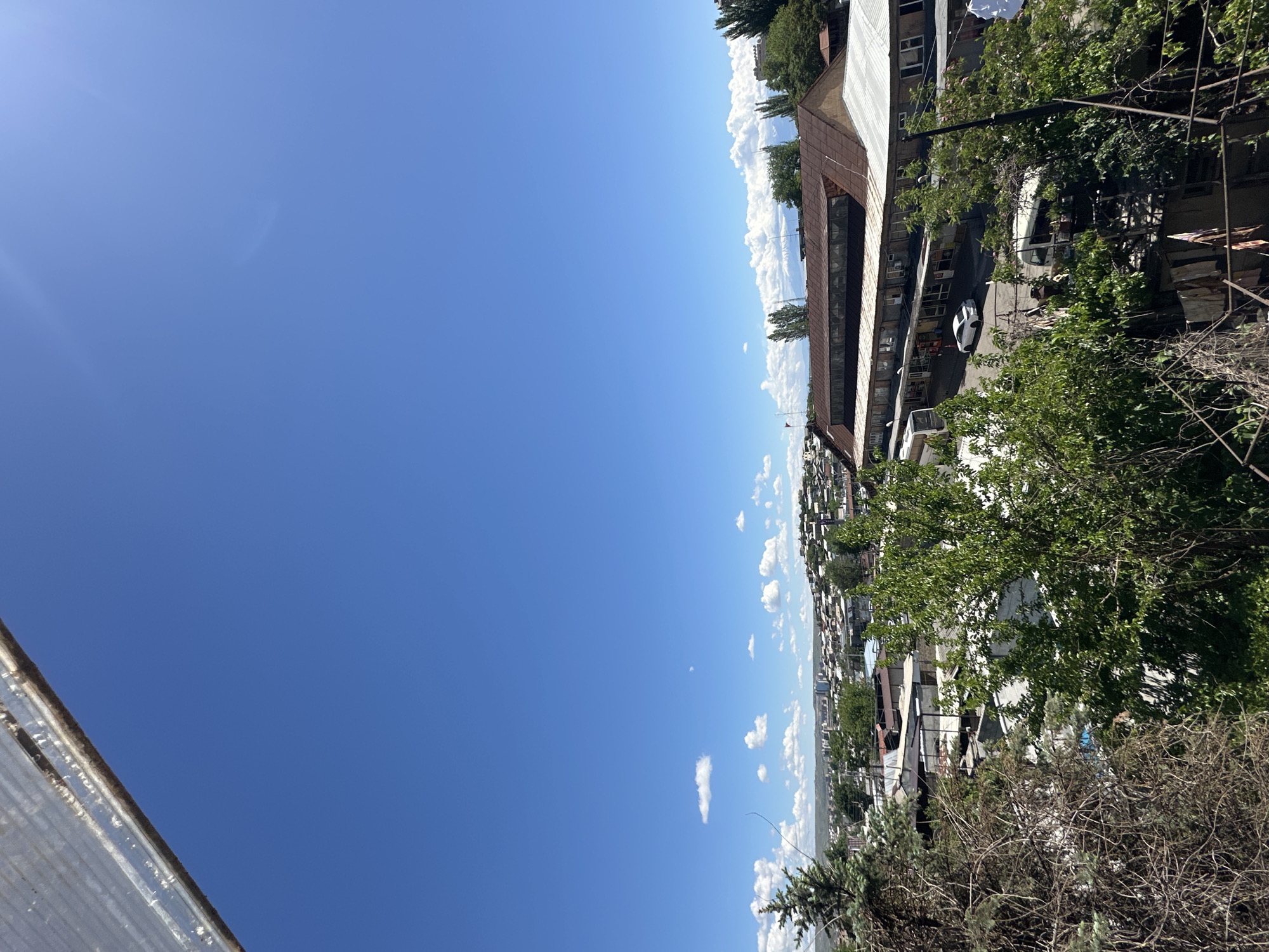 The image depicts a daytime scene in Yerevan, Armenia, showcasing a cityscape under a clear blue sky with scattered clouds. A multi-story building with a rusted roof and multiple windows is visible, with a white car parked in its vicinity. Trees and dense foliage partially obscure the view, indicating a residential or urban environment. The image's perspective is taken from an elevated position, looking down upon the scene.