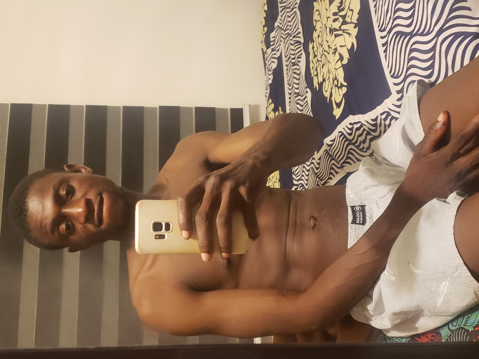 A man is taking a selfie indoors. He is lying on a bed with a patterned headboard and bedspread. The location is likely a residence in Adonkolo, Nigeria. He is shirtless and wearing shorts. He is holding a gold-colored smartphone.