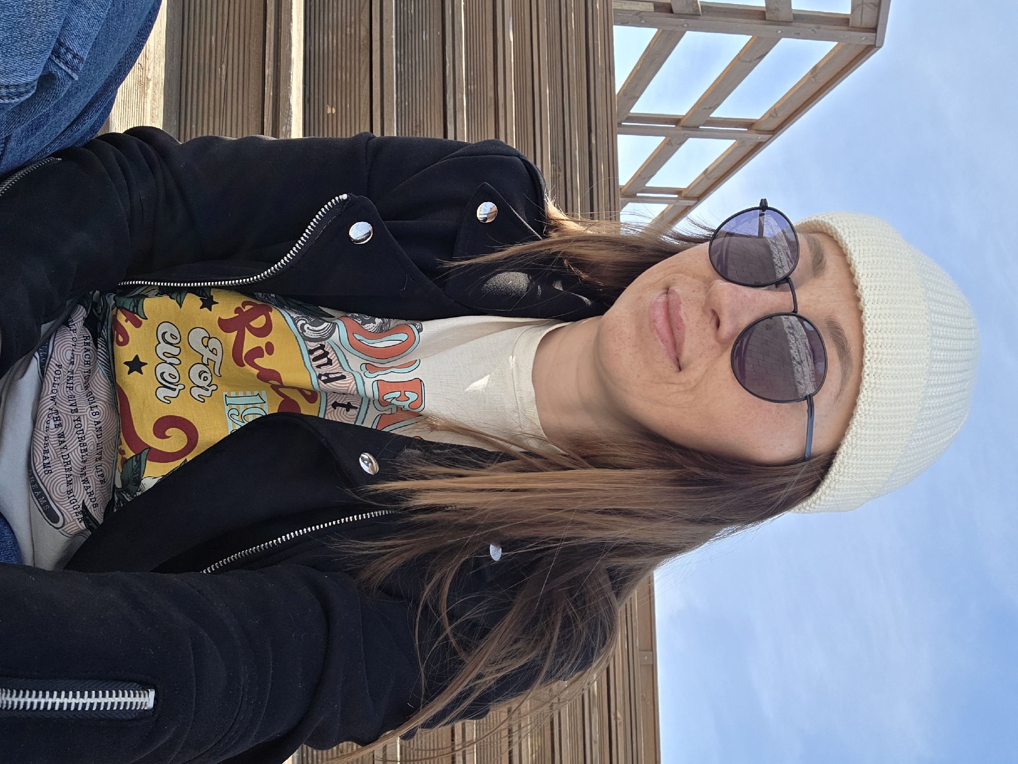 A woman is depicted in a medium close-up, seated outdoors on a tiered wooden structure in Votkinsk, Russia. She is positioned slightly to the viewer's right, with her gaze directed generally forward. She wears a cream-colored ribbed knit beanie, dark round sunglasses, and a black jacket with silver snaps on the lapels and a visible silver zipper. Beneath the jacket, a light-colored graphic t-shirt with colorful text, including "For ever," is visible. The lower left of the frame shows a portion of blue jeans. Her long, brown hair falls over her shoulders. Freckles are discernible on her nose and cheeks, and her lips are slightly parted. The background features a wooden railing and a clear, bright blue sky. The scene is illuminated by natural daylight.
