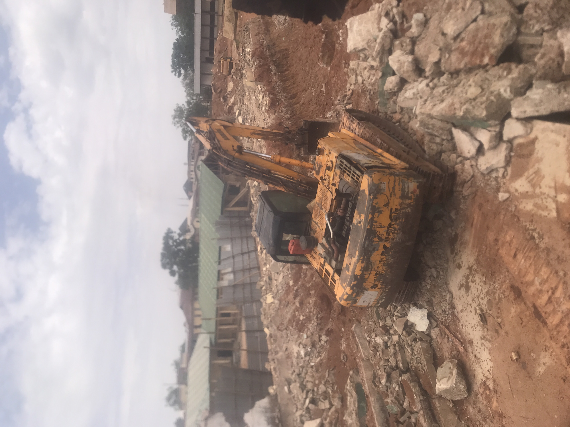 The image shows an excavator on a construction site in Jos, Nigeria. The excavator is yellow and appears to be in use, surrounded by piles of rocks and excavated earth. A building under construction can be seen in the background, indicating active construction work. The sky is overcast, suggesting a cloudy day.