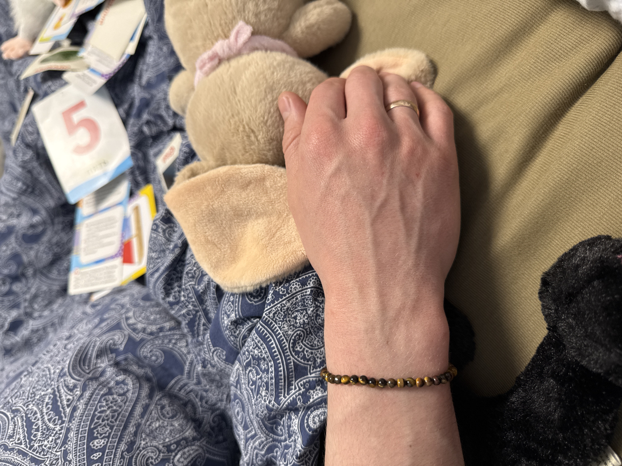 A person's hand, adorned with a gold wedding band and a beaded bracelet, rests on a tan stuffed bunny. The bunny, with a pink bow, sits on a blue paisley-patterned surface. Scattered nearby are children's learning cards, one prominently displaying the number "5". A brown upholstered surface is visible to the right. The image does not provide information on Moscow, Russia.