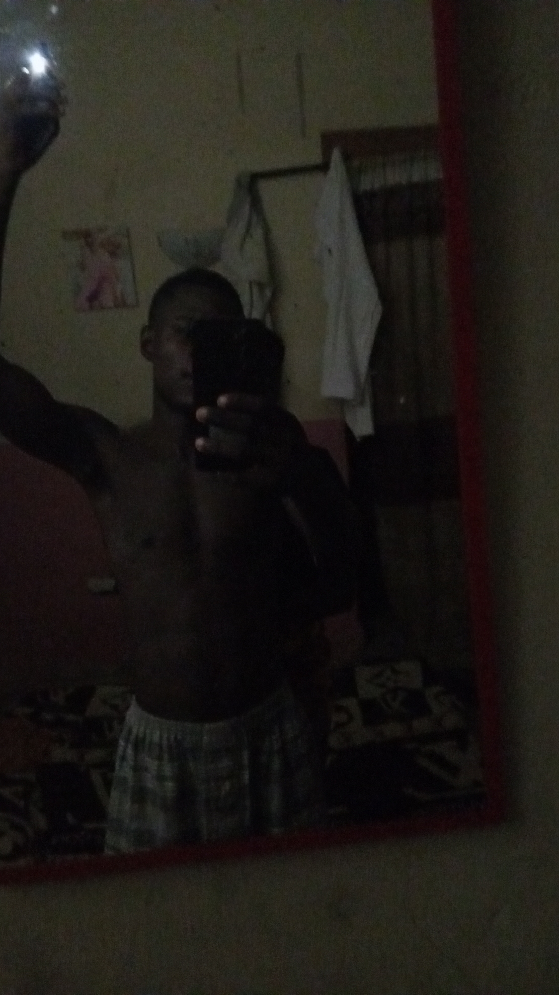 A person is taking a mirror selfie in what appears to be a bedroom. The person is shirtless and wearing checkered shorts. The room is dimly lit, and the background shows a wall with some framed pictures, a window with blinds, and some clothing hanging on a rack. The overall impression is of a casual, personal space. The presence of a framed picture of a person in traditional attire suggests a possible cultural context, and if the user's request is to situate it in Jalingo, Nigeria, then the image would be depicting a scene from that city.
