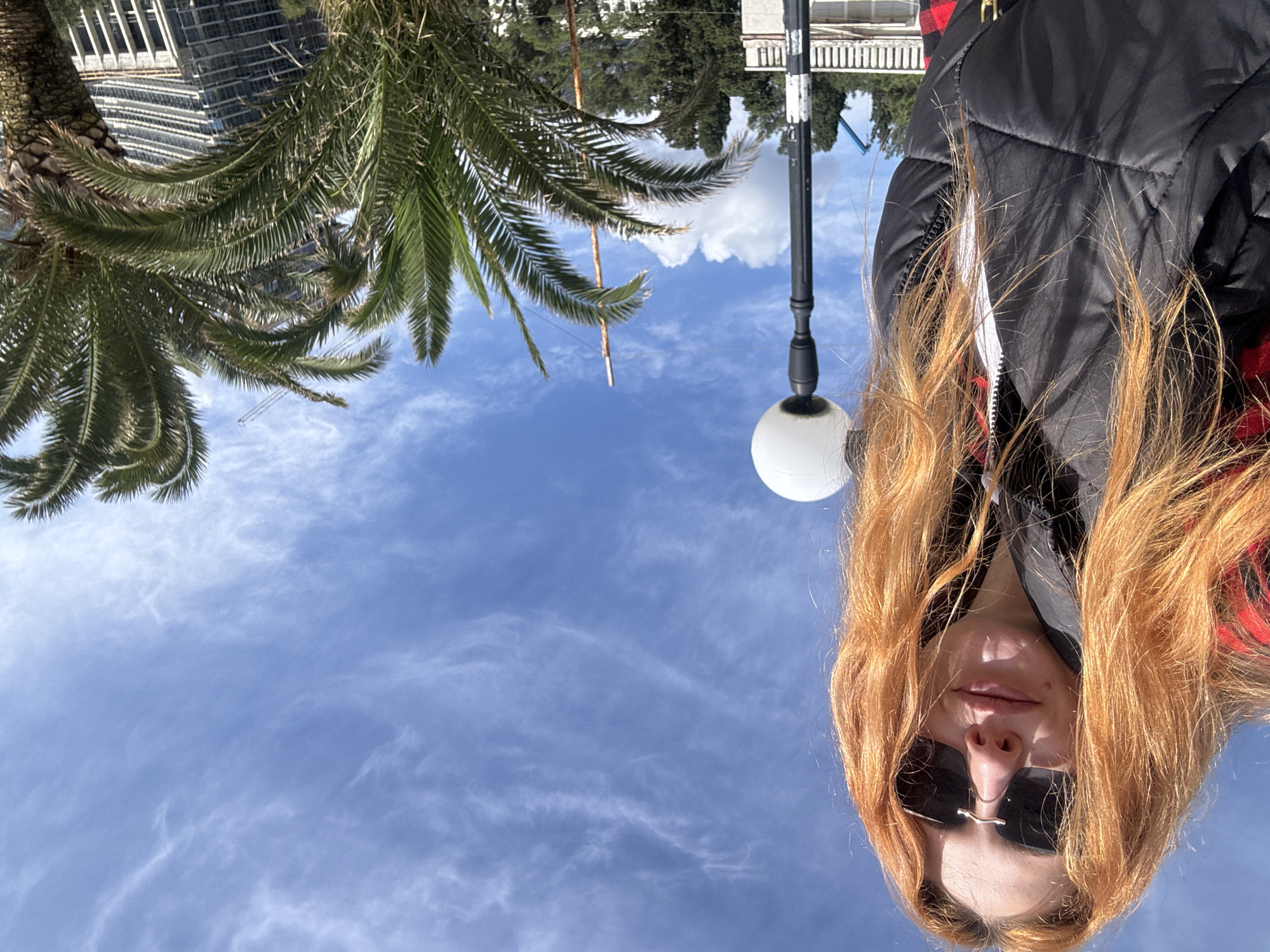 An outdoor portrait shot in Budva, Montenegro, captures a person from a low angle, looking upwards towards a vibrant blue sky. In the foreground, positioned centrally, is a young person with long, flowing reddish-blonde hair, wearing black sunglasses that obscure her eyes. She is dressed in a black puffer jacket, with hints of a red and black plaid shirt visible underneath.
To the left of the person, a tall black street lamp with a spherical white light fixture extends vertically into the frame. In the background, on the upper left side, a large palm tree with lush green fronds adds a tropical element to the scene. Beyond the palm tree, part of a modern building with a glass facade is visible, suggesting an urban setting. The upper portion of the image is dominated by a clear, bright blue sky with scattered, wispy white clouds. The bright daylight indicates it is daytime. No specific activity beyond posing for the photograph is depicted, and no text is visible in the image.