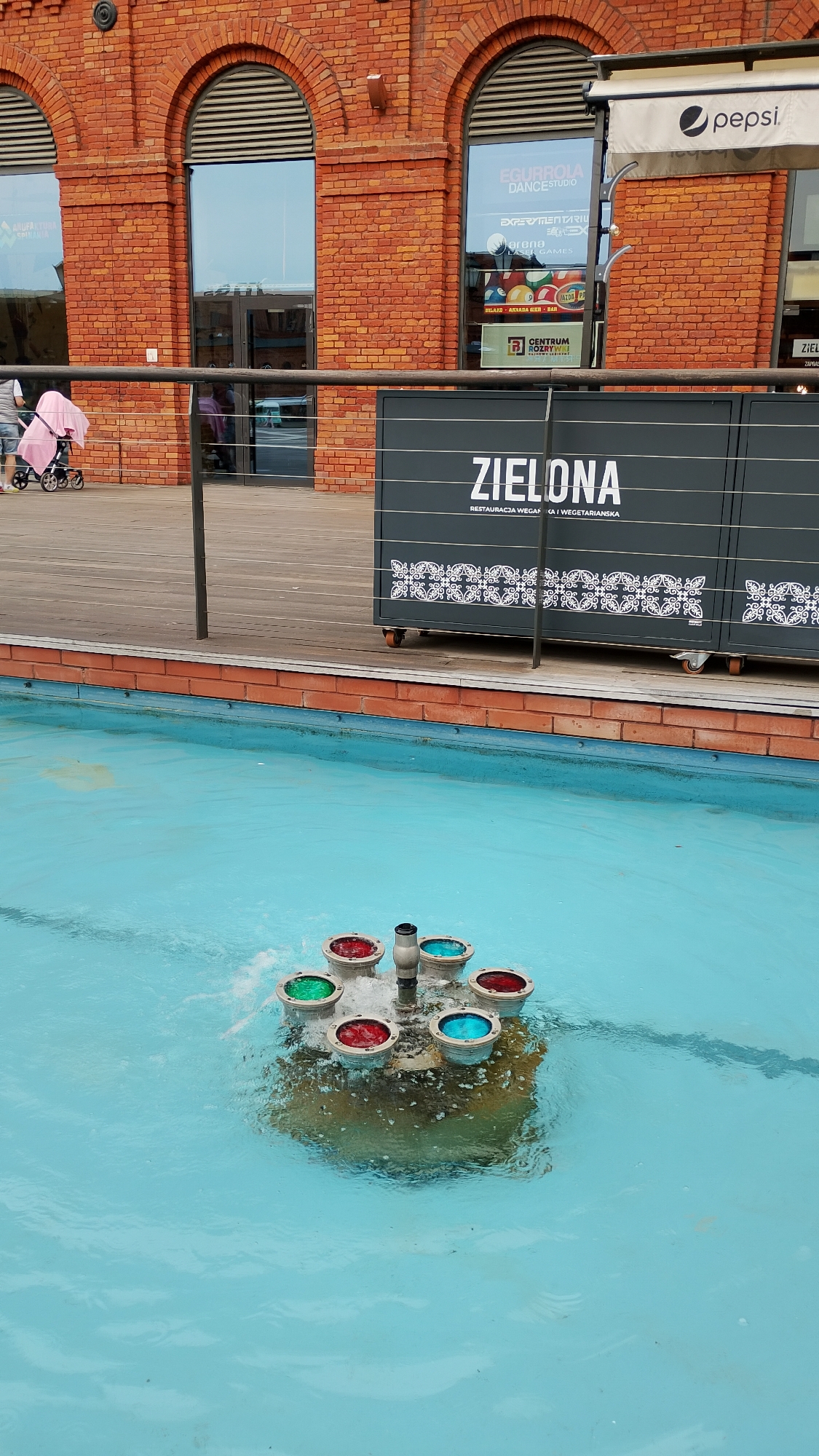 The image depicts a water fountain in the Manufaktura shopping and entertainment complex in Łódź, Poland. The fountain has a central structure with several circular containers around it, lit up with green, red, and blue lights. The water in the fountain is a light blue color.
In the background, there is a brick building with arched windows. A black metal railing runs along the edge of a wooden platform. Behind the railing, a sign reads "ZIELONA," with the words "RESTAURACJA WEGAN" underneath. A Pepsi sign is visible above one of the building entrances.
A person pushing a stroller is faintly visible in the background on the left side of the image.