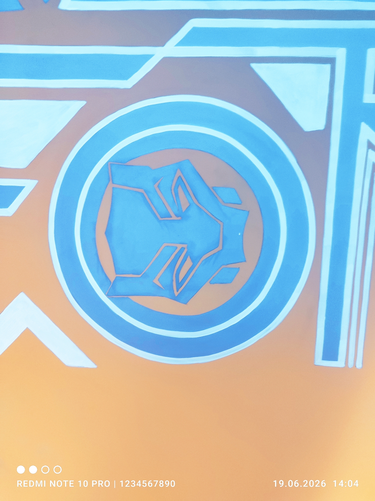 The image shows a close-up of a mural with abstract geometric shapes in blue and white against an orange background. In the center is a circular emblem with a stylized blue fist-like shape inside, outlined in a darker blue. The mural appears to be painted on a flat surface. The bottom of the image contains text overlays: "REDMI NOTE 10 PRO | 1234567890" on the left, and "19.06.2026 14:04" on the right. There are also four white circular dots arranged horizontally on the left side of the text. The mural's design suggests it might be related to a sports team, a logo, or an artistic representation of strength or unity. There is no visible context to determine its specific location within Fergana City, Uzbekistan.