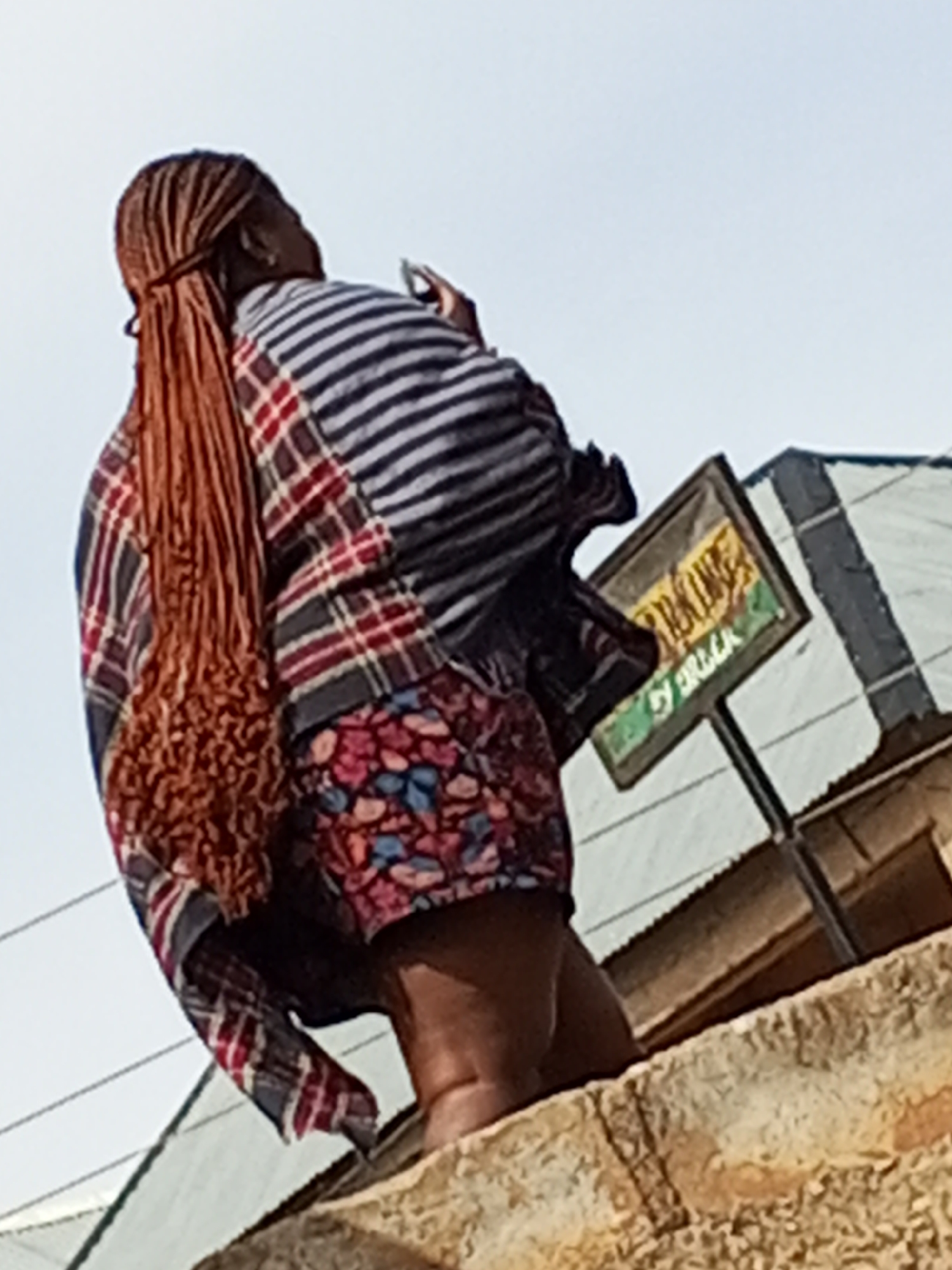 An eye-level shot captures a female individual in Gwong, Nigeria, standing outdoors on a masonry structure. She is facing away from the camera. The woman has long, reddish-brown braided hair that extends down her back. She is wearing a multicolored outfit consisting of striped and plaid patterns. On the lower part of her body, she is wearing patterned shorts. She appears to be holding a small device, possibly a phone, in her right hand.
Behind her, a weathered sign with visible text is mounted on a pole. Corrugated metal roofs of nearby structures are also visible in the background. The sky is a pale blue, suggesting daylight conditions. The overall environment seems to be urban and possibly a neighborhood in Gwong.