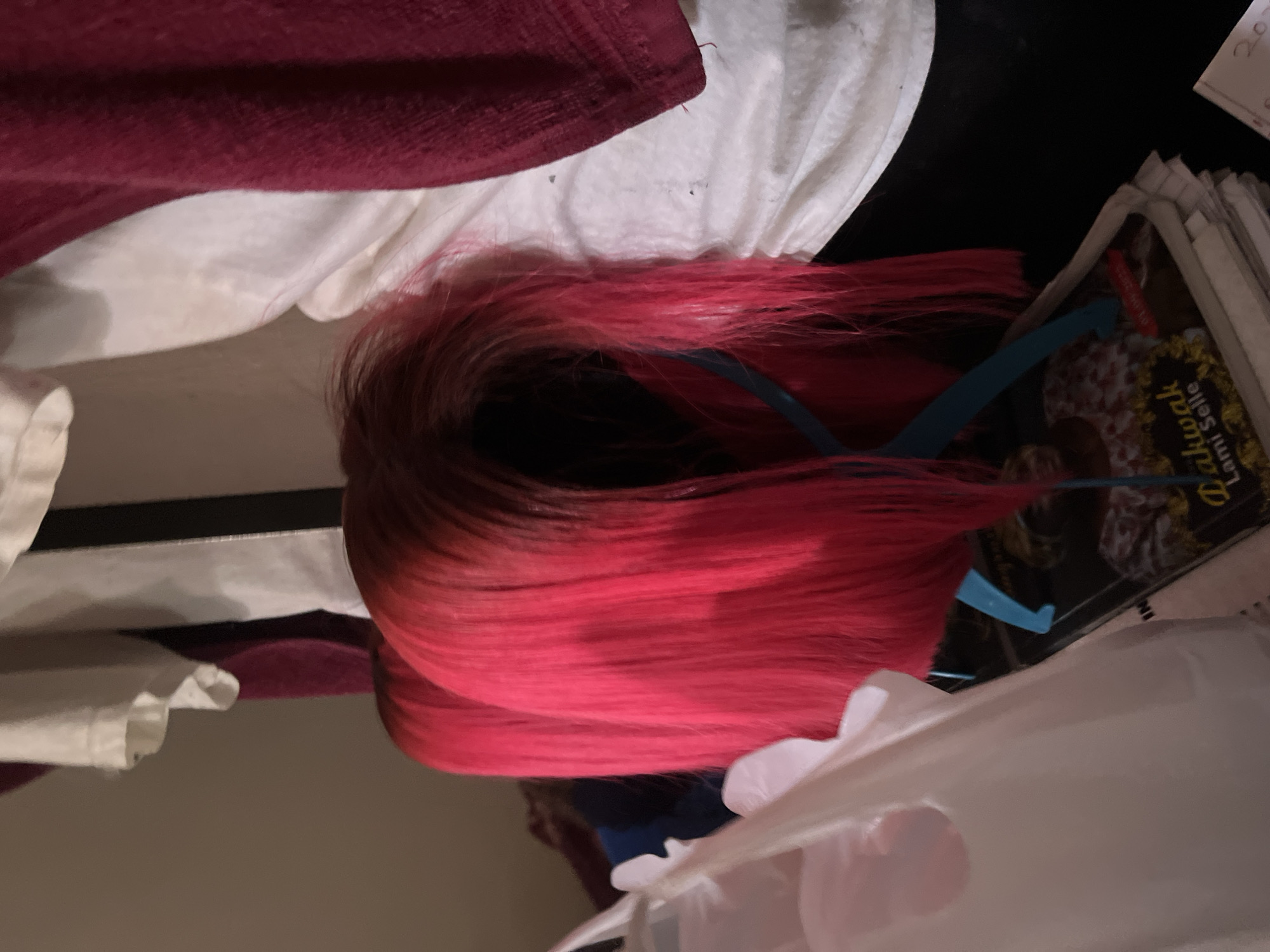 A vibrant pink wig rests on a stand in a cluttered indoor setting, possibly a bedroom or closet, in Jos, Nigeria. The wig has a gradient color, transitioning from a darker shade at the roots to a bright pink at the ends. It is styled straight and appears to be synthetic. In the background, various fabrics are visible, including a white t-shirt and a dark red towel or cloth. A white plastic bag is partially in the foreground to the left. Below the wig stand, a stack of magazines or papers is visible, with some text and images discernible on the top pages, including what appears to be a person's face and the words "Dakiwak Lami Selle". The lighting is dim, suggesting it may be evening or the room is not well-lit. The overall impression is one of personal belongings and preparations for styling or display.