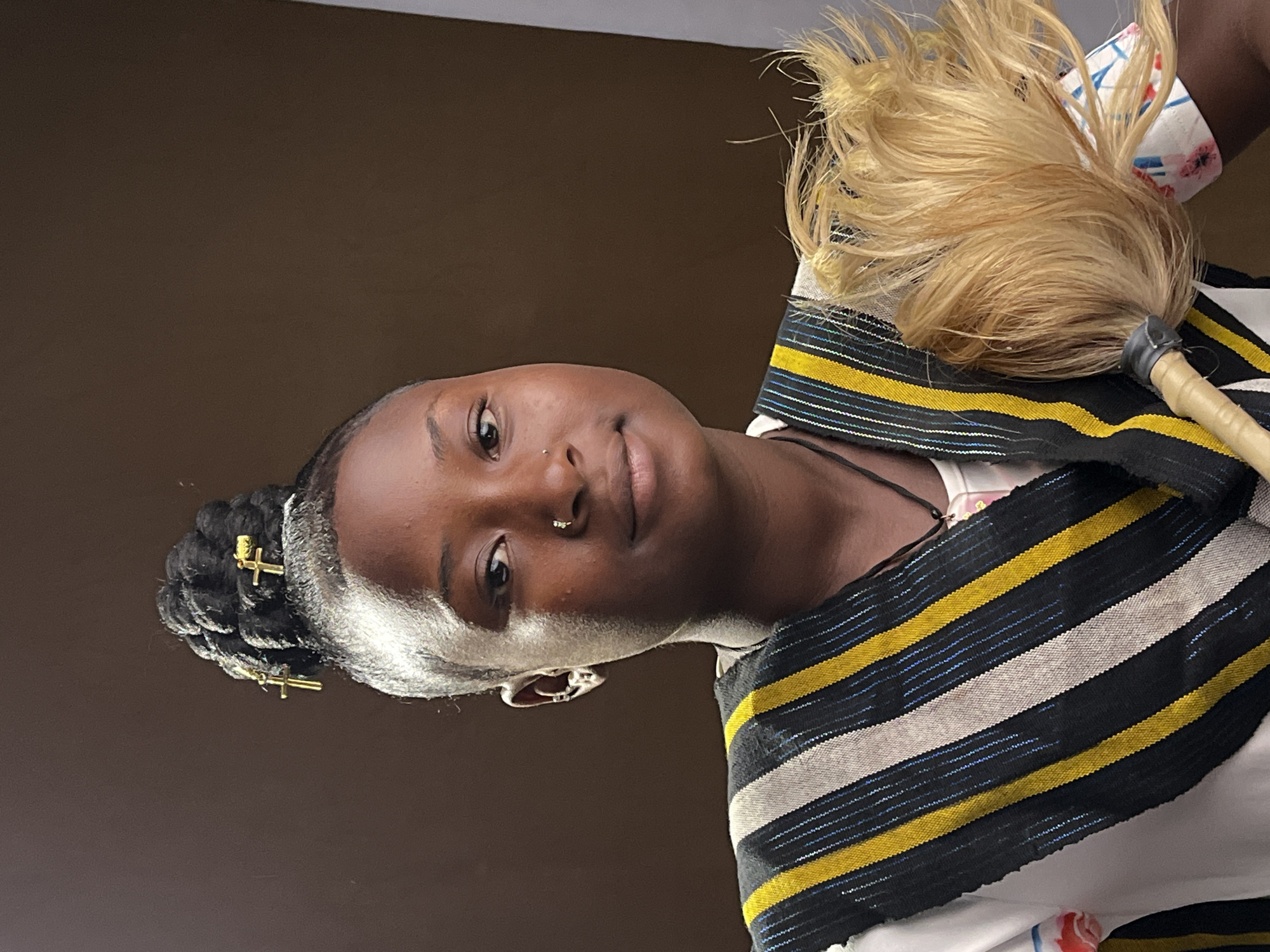 The image features a portrait of a young Black female. She is looking directly at the viewer with a gentle, soft smile.
Her hair is styled in dark braids, with some lighter (possibly white or very light blonde/grey) strands visible at the front and sides of her head. Several small, gold-colored, cross-shaped hair clips are embedded in her braids on the left side of the frame. She has a small stud nose piercing on her left nostril.
She is wearing a vibrant striped garment, possibly draped over her shoulders or chest. The fabric features horizontal stripes in black, yellow, and a lighter color (appearing white or light grey), interspersed with thin blue lines. A plain white undergarment and a thin black strap are visible beneath the striped fabric.
On the right side of the image, a decorative object is visible, held or placed near her. It consists of light blonde, feathery material attached to a light wooden handle with a dark grey or black cap. This object resembles a traditional fly whisk or a ceremonial fan.
The background is a plain, solid dark brown wall, suggesting an indoor setting. The overall expression of the person is calm and pleasant. Given the provided context, the image likely depicts a individual in Jos, Nigeria, potentially showcasing elements of cultural attire or accessories. There is no visible text in the image.