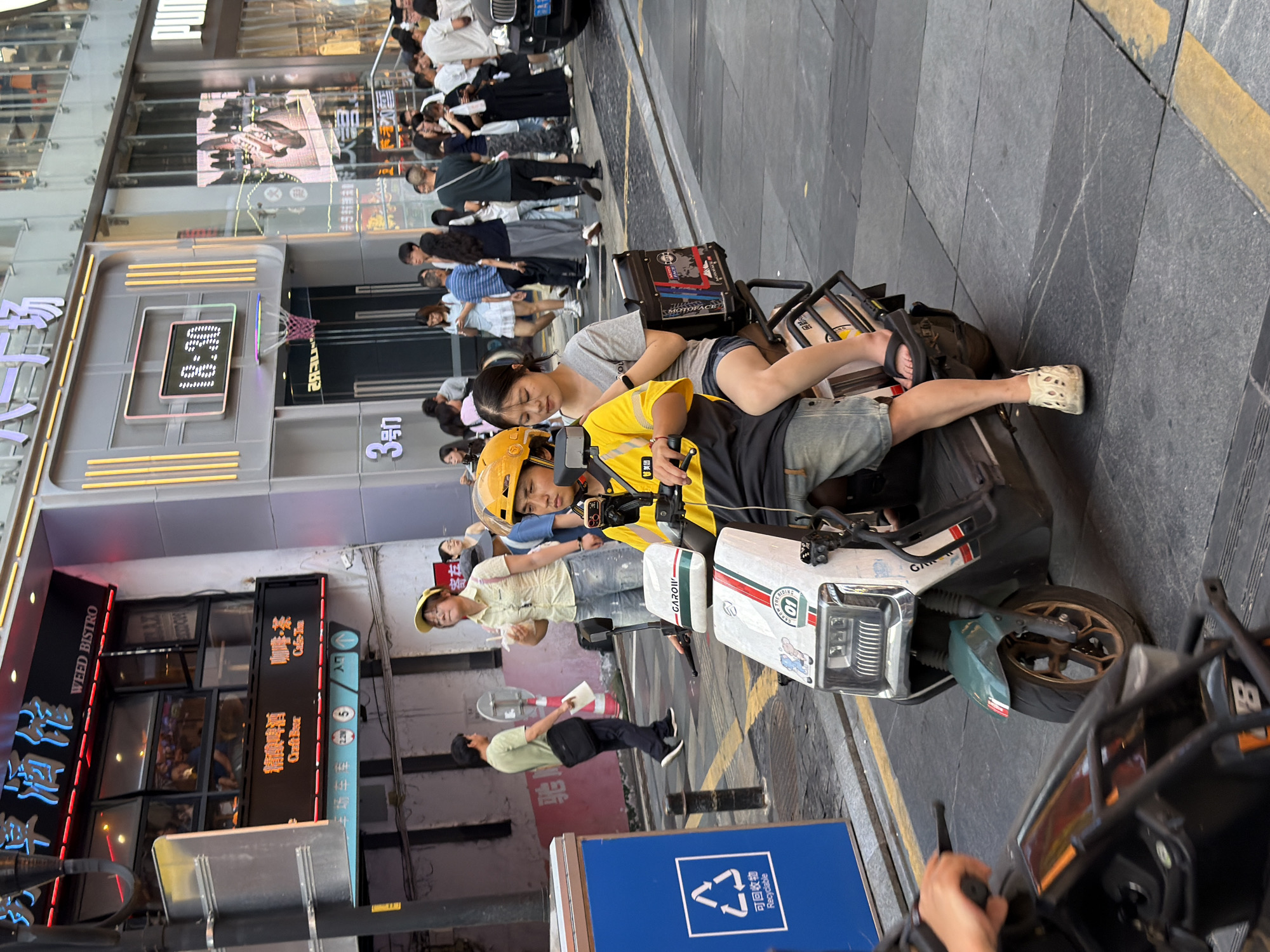 The image shows a bustling street scene in Yuzhong District, China, with a focus on two individuals riding a scooter. A young woman, wearing a light-colored shirt and denim shorts, sits behind the driver, who is wearing a yellow shirt and helmet. Both appear to be casually dressed. The scooter is white with green accents and has the text "MOTORACE" and "GAROII" visible on its body. It also has a black storage box on the back.
In the background, several other people are visible, suggesting a busy urban environment. Storefronts with neon signs line the street. One sign reads "WEED BISTRO" and "精酿啤酒 Craft Beer," indicating a food and beverage establishment. Another sign says "咖啡·茶 Cafe-Tou." There are also signs with Chinese characters, one of which appears to be "3号" (No. 3). A blue recycling bin with Chinese text and the recycling symbol is also present.
The image is taken from a slightly low angle, looking up towards the subjects on the scooter. The lighting suggests it is daytime, possibly late afternoon or early evening, given the shadows and the brightness of the signs. The overall atmosphere is lively and characteristic of a busy commercial street in a Chinese city. The people appear relaxed and engaged in their activity, with no obvious signs of distress or urgency. The presence of the scooter and the storage box implies they might be on an errand or delivery.
