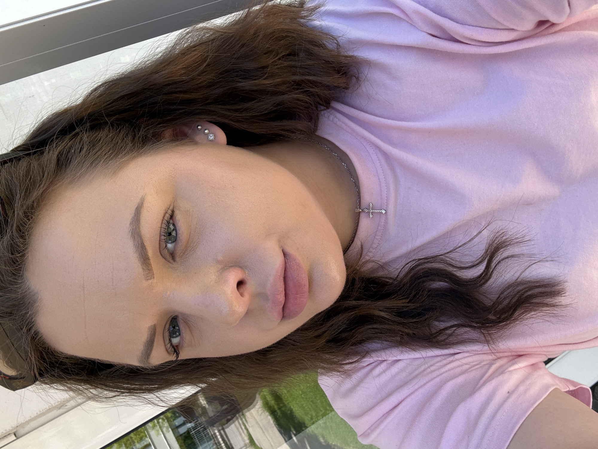 A light-skinned woman with brown hair and light green eyes is positioned in a sideways portrait. She wears a light pink t-shirt and a silver chain necklace with a small cross pendant. She has piercings in her ear. The background includes part of a building and greenery, and the photo was taken outdoors during the daytime. While not explicitly confirmed, the prompt mentions the city of Lovech, Bulgaria.