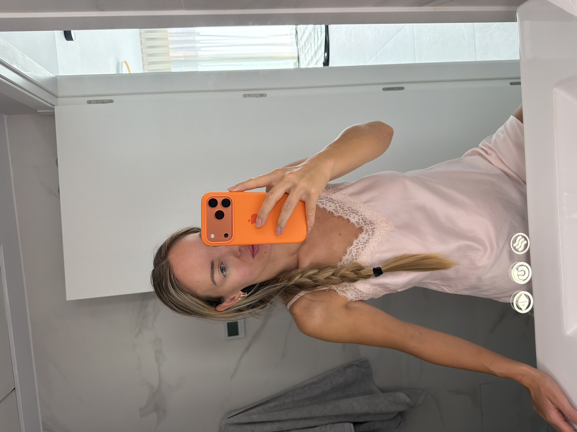 A fair-skinned woman with blonde hair in a braid takes a mirror selfie in a modern bathroom. She is wearing a pale pink slip with lace trim. She holds an orange-cased smartphone in her right hand to take the photo. The bathroom has marble-patterned walls and a white vanity with three illuminated circular controls. A gray towel hangs on the left, and a white door is visible on the right, revealing a glimpse of a room with sheer curtains. The scene is likely in Finestrat, Spain, suggested by the query, though this cannot be definitively confirmed from the image alone.