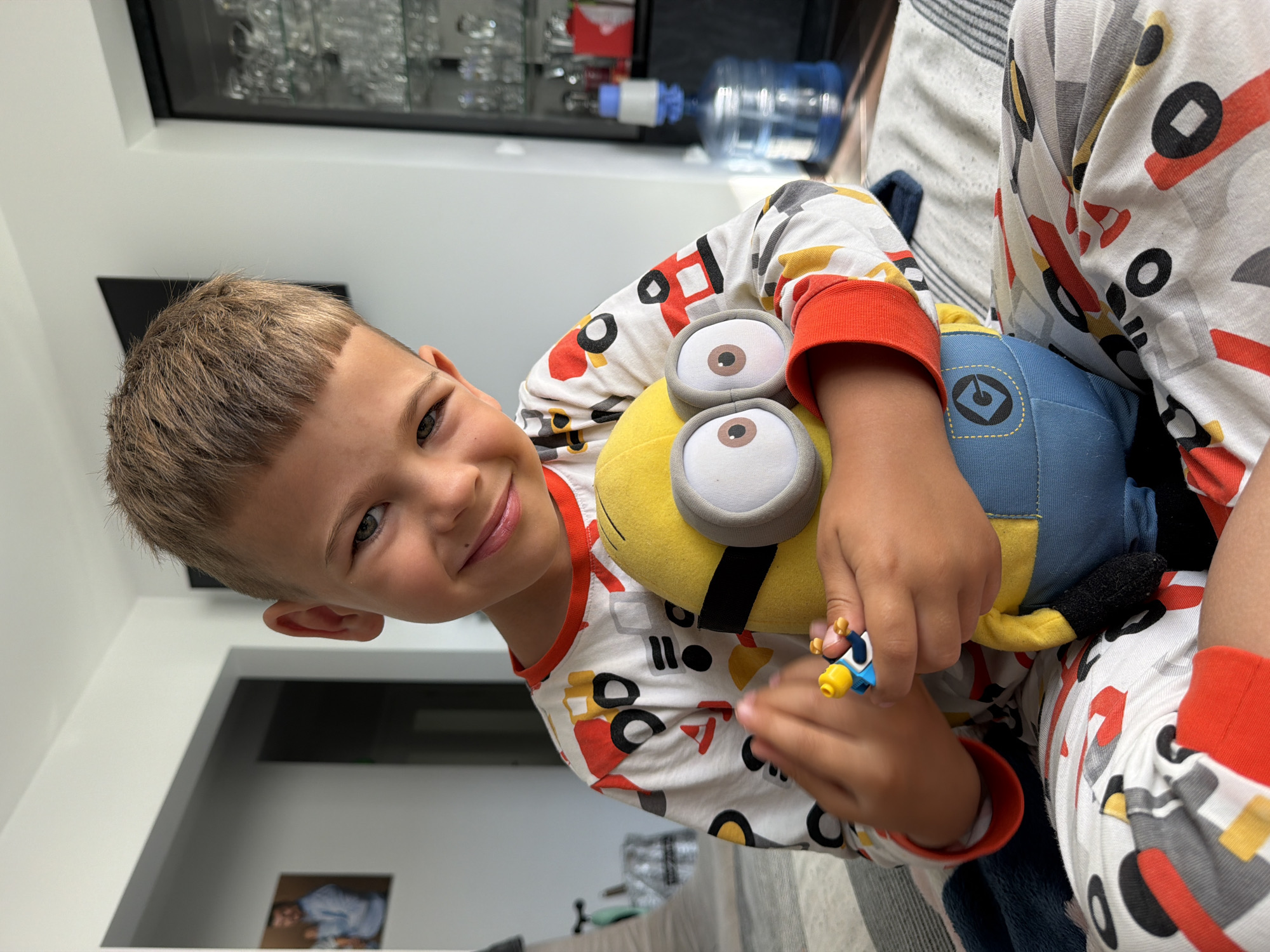 A young boy with short, light brown hair and light skin is seated indoors, looking at the camera with a slight smile. He is wearing pajamas with a pattern of construction vehicles. He is holding a yellow stuffed Minion toy and a small blue and yellow figure in his hands. The background shows a room with a white wall, a framed picture on the wall, and a glass cabinet with various items inside. There is also a large blue water bottle with a dispenser in the background.
