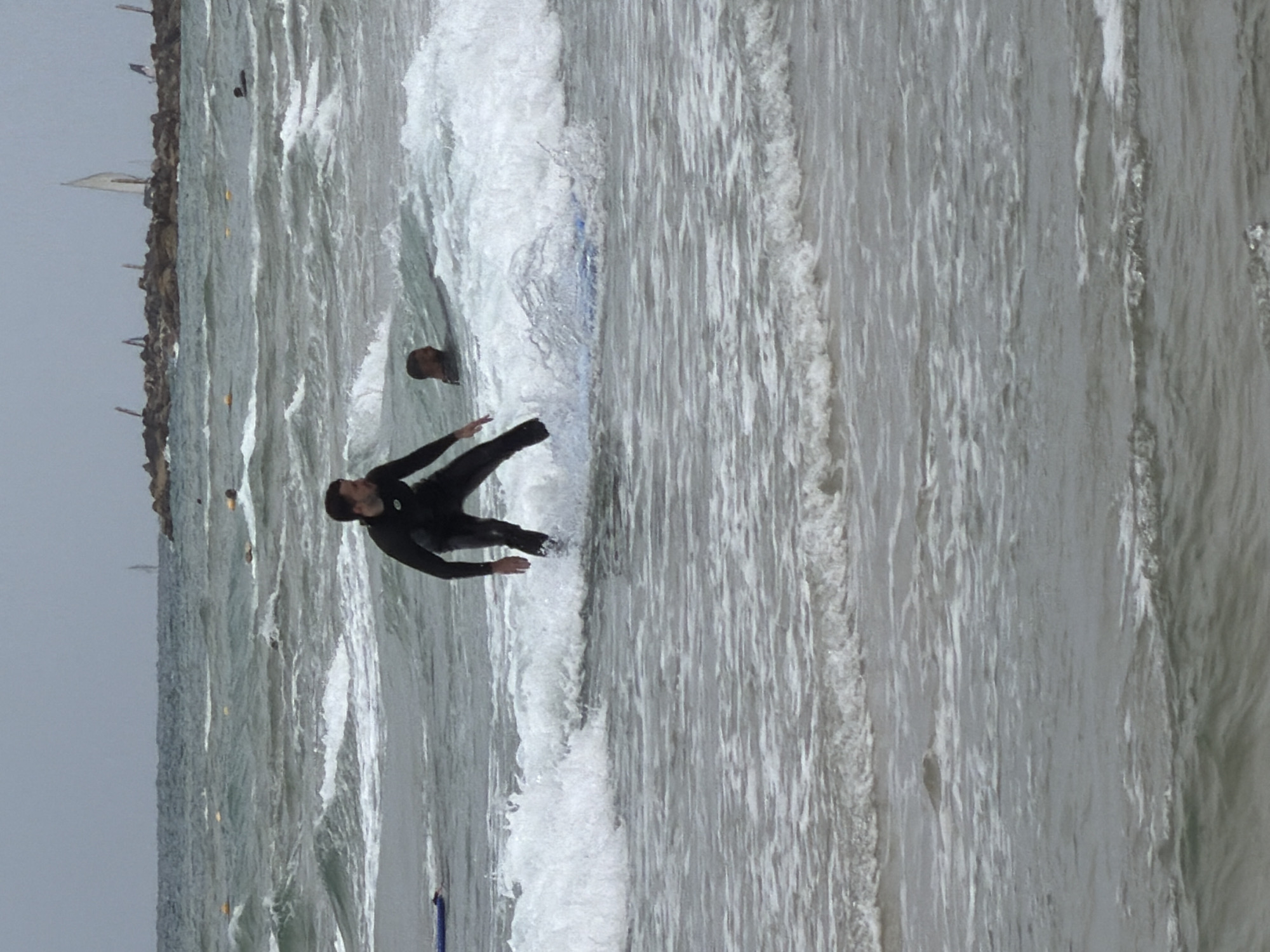 A man in a black wetsuit is riding a wave on a surfboard in the Mediterranean Sea. He is positioned on the left side of the frame. To his left is a head poking out of the water, and further to the left is the Tel Aviv coastline. The sky is overcast.