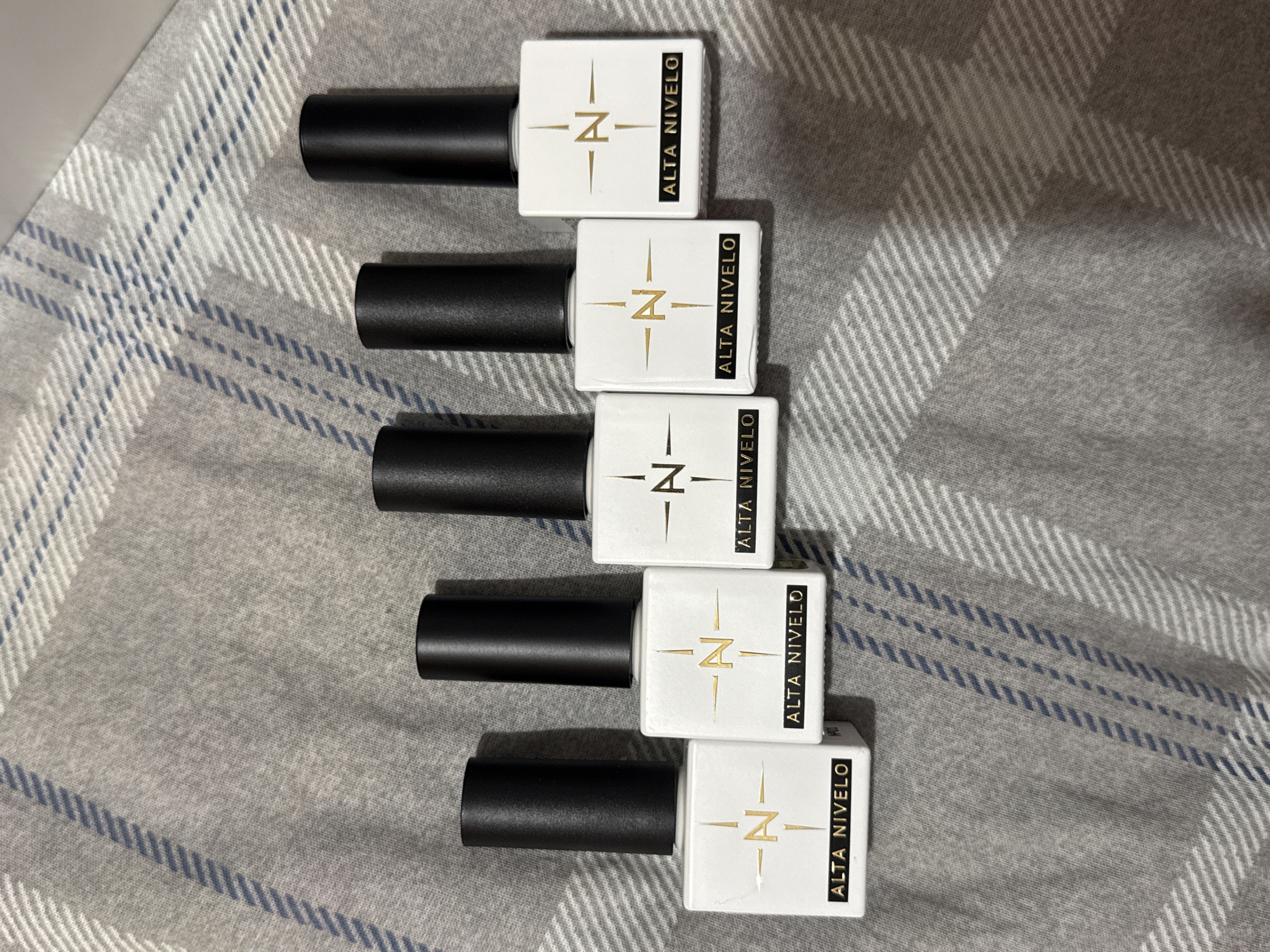 Five bottles of nail polish are arranged in a diagonal line on a grey and white plaid fabric surface. Each bottle has a white square base with a black cap. On the front of each bottle, there is a gold logo consisting of a stylized "N" with radiating lines, and below the logo, the text "ALTA NIVELO" is printed in gold. The bottles are positioned to face the viewer. The fabric surface appears to be a blanket or throw, with a repeating plaid pattern of grey and white lines. There are no people or other objects visible in the image. The lighting suggests an indoor setting. The location is not discernible from the image itself.