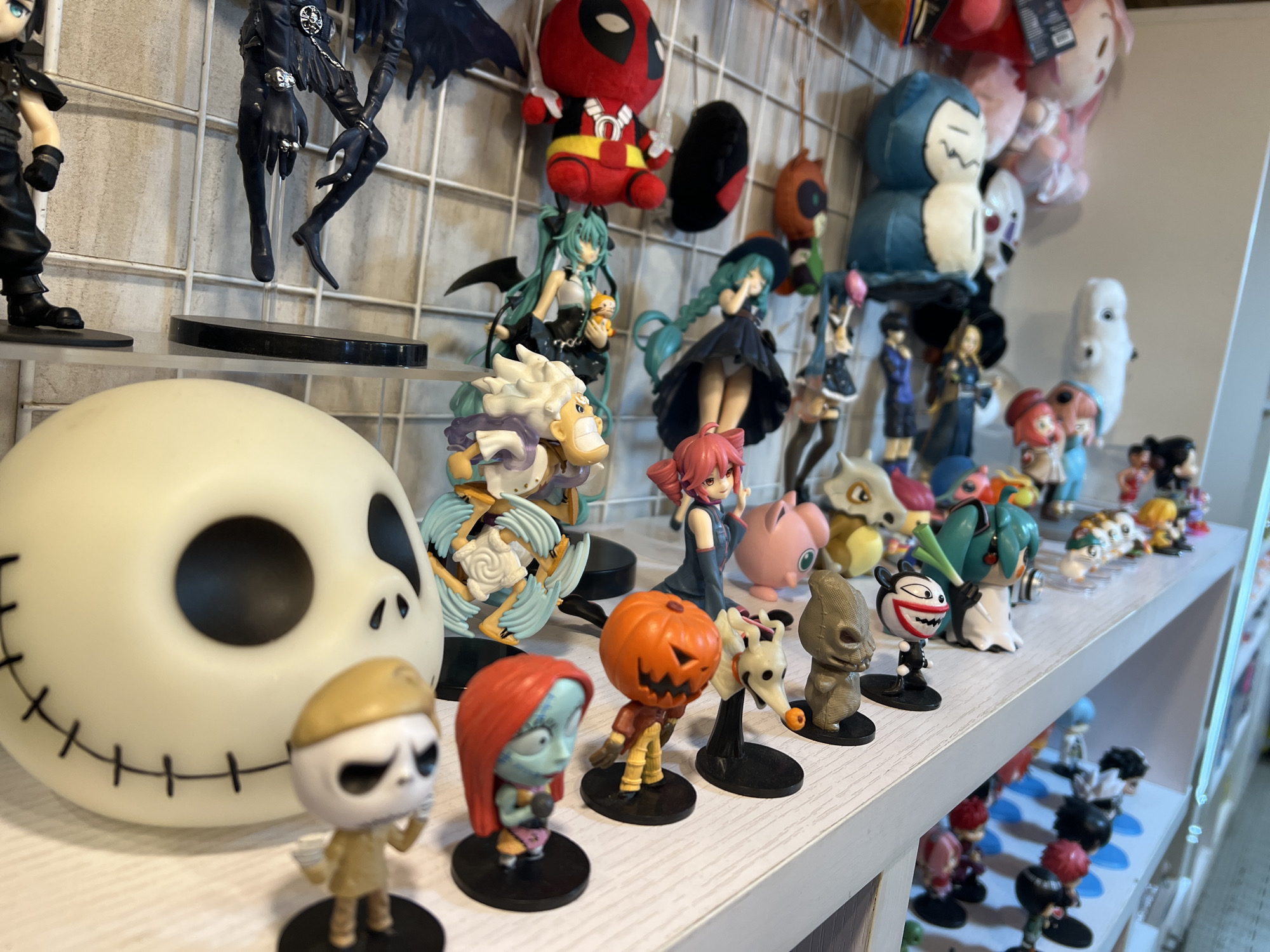 The image shows a shelf filled with various anime figurines and plush toys. The figurines are arranged in rows, with some larger items like a Jack Skellington head in the foreground. The background consists of a grid-like wall and a white shelving unit. The lighting suggests an indoor setting, possibly a retail store or a personal collection display. The specific location in Arica, Chile, is not discernible from the visual information provided in the image.