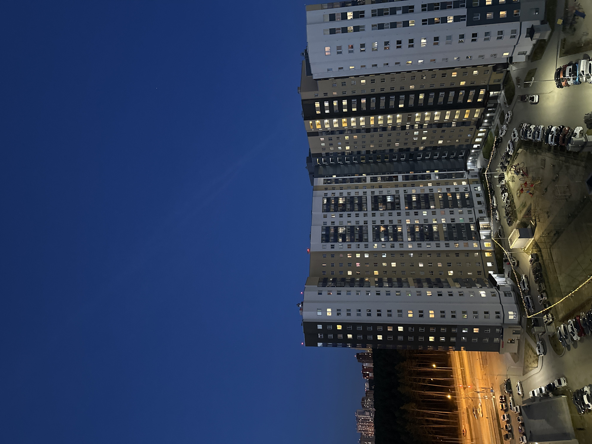 This vertical image captures an urban evening or night scene in Yekaterinburg, Russia, dominated by a towering residential building against a deep blue sky. The multi-story building, stretching from the top to the bottom of the right side of the frame, features a modern design with sections of light-colored (white/grey) and darker (beige/black) facades. Numerous windows are visible across its many floors, with a significant number of them brightly illuminated, indicating occupancy and the late hour.
At the base of the building, a well-lit ground area unfolds, including a large parking lot densely filled with various cars. Adjacent to the parking, sections of a children's playground, complete with slides and other equipment, are visible under artificial light. Further right and extending into the background, a wide road or highway is illuminated by rows of streetlights, creating light streaks that suggest ongoing vehicle movement. In the far distance, the faint glow of other city lights and buildings can be discerned.
The left side of the frame is dominated by a clear, dark blue sky, indicative of dusk or nighttime, with a single, very faint point of light visible, possibly a star or distant aircraft. The weather appears clear, with no clouds. The scene conveys the quiet activity of a modern residential complex after sunset, illuminated by a network of artificial lights. No discernible text is visible in the image.