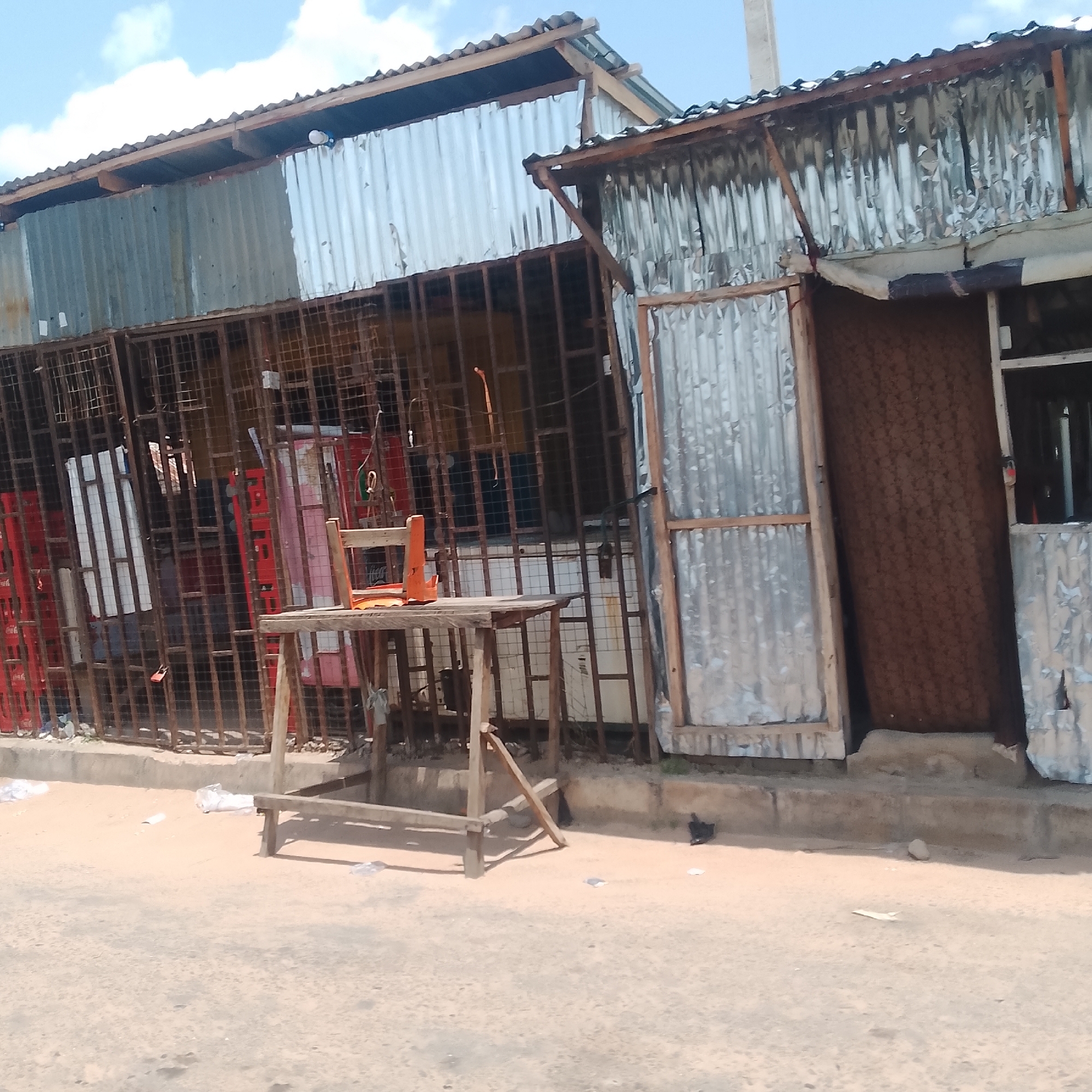 The image depicts an outdoor scene in Jalingo, Nigeria, featuring a makeshift market stall constructed with corrugated metal sheeting and metal bars. In the foreground, a wooden table with two simple wooden chairs sits on a dirt ground. Behind the table, the market stall is partially visible, showcasing red crates and what appear to be refrigerators or coolers. A weathered metal cage-like structure forms the front of the stall, with additional corrugated metal and wooden elements making up the sides and roof. The environment suggests a commercial area, likely a street or roadside market, with debris scattered on the ground. The sky above is partly cloudy.