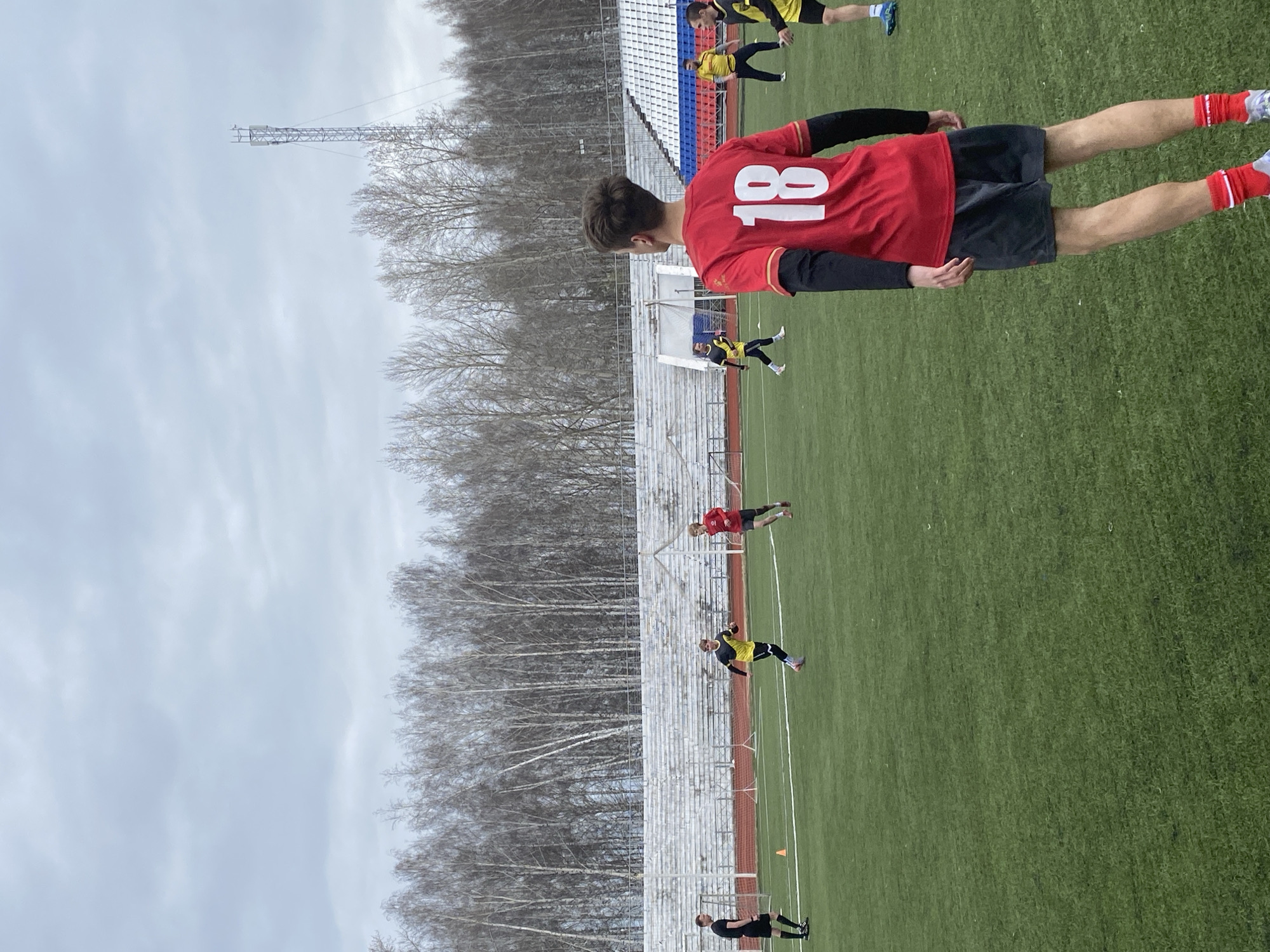 The image depicts a soccer field in what appears to be Chaikovsky, Russia, based on architectural styles. It shows a man in a red jersey with the number 18 on the back. He is standing in the foreground. Other people are practicing on the field, dressed in soccer attire. A set of metal bleachers can be seen behind them. In the background, there is a bare forest and a communication tower under an overcast sky. The field is green and the ground is covered in grass.