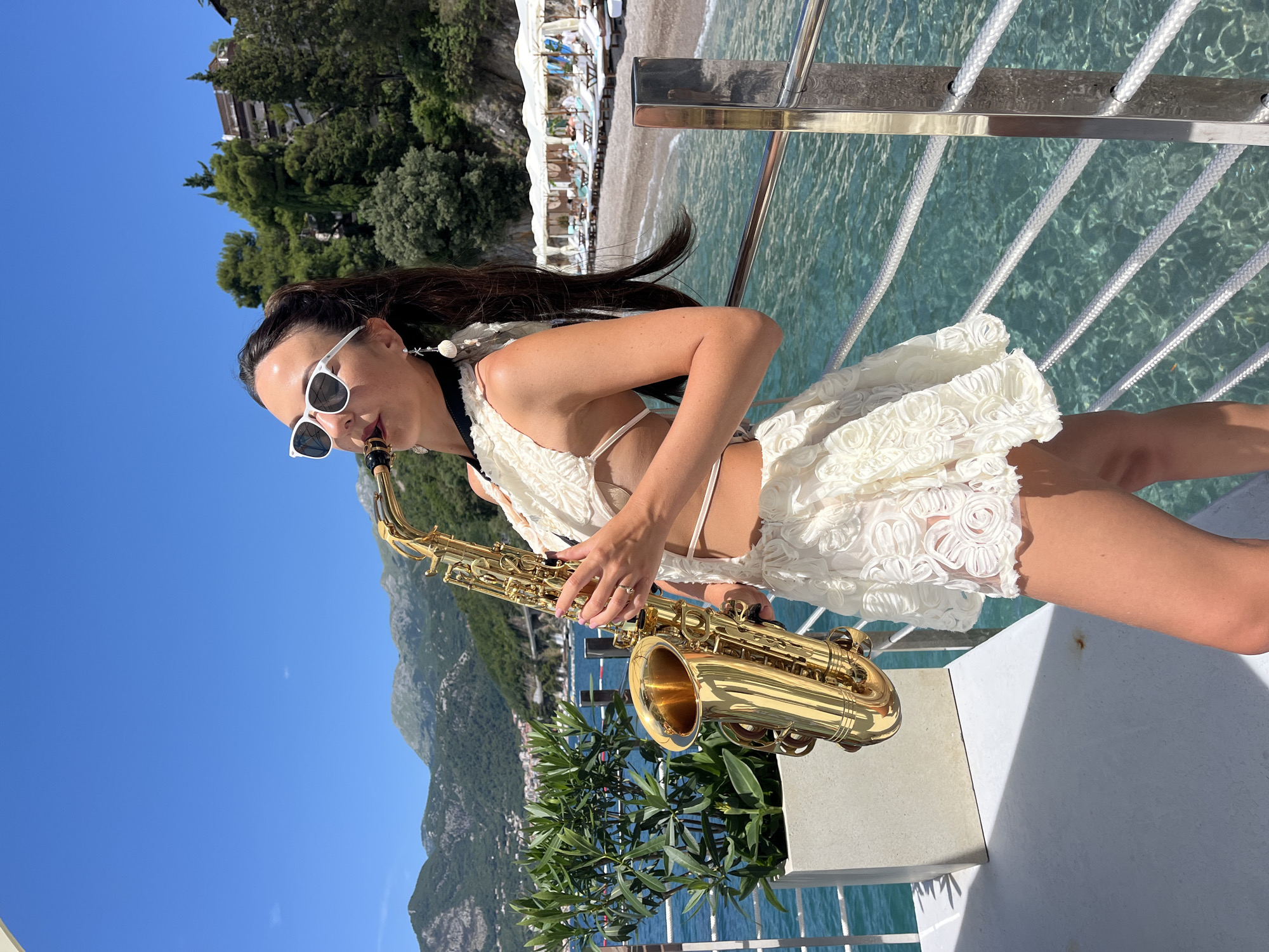 A woman is playing a saxophone on a boat in front of a beach in Budva, Montenegro. The woman is wearing a white lace outfit and sunglasses. The beach has lounge chairs and umbrellas. The water is clear and blue, and the sky is bright and sunny. Mountains are visible in the background.