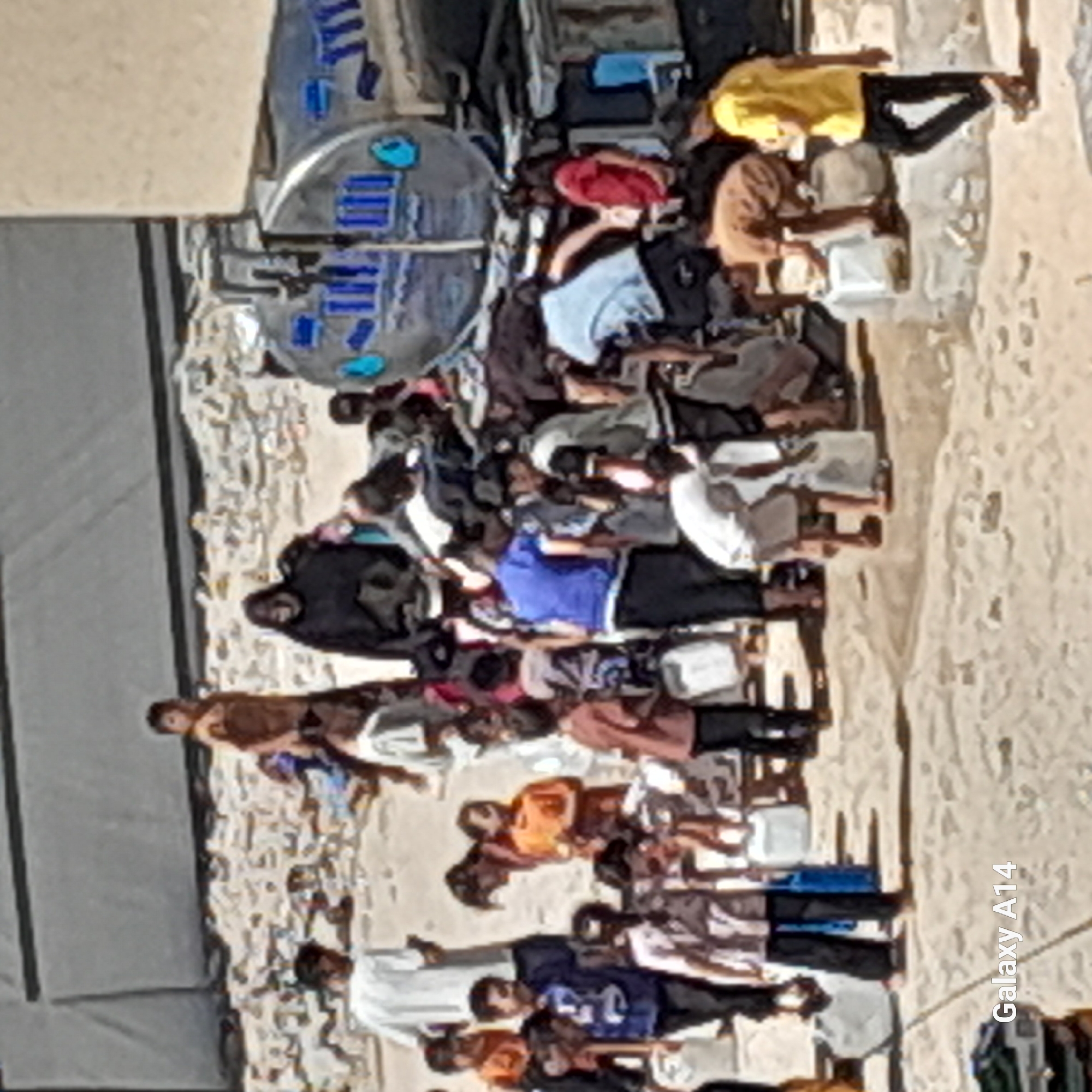 In the city of Khan Yunis, Palestinian Territories, a group of people, including men, women, and children, are gathered in an outdoor area. The ground is covered with rubble and debris, suggesting a damaged environment. Some individuals are carrying or interacting with containers, potentially collecting or distributing resources. A silver water truck with blue markings is present, indicating a possible aid or water supply operation. The scene appears to depict a community dealing with challenging circumstances, possibly related to displacement or scarcity.