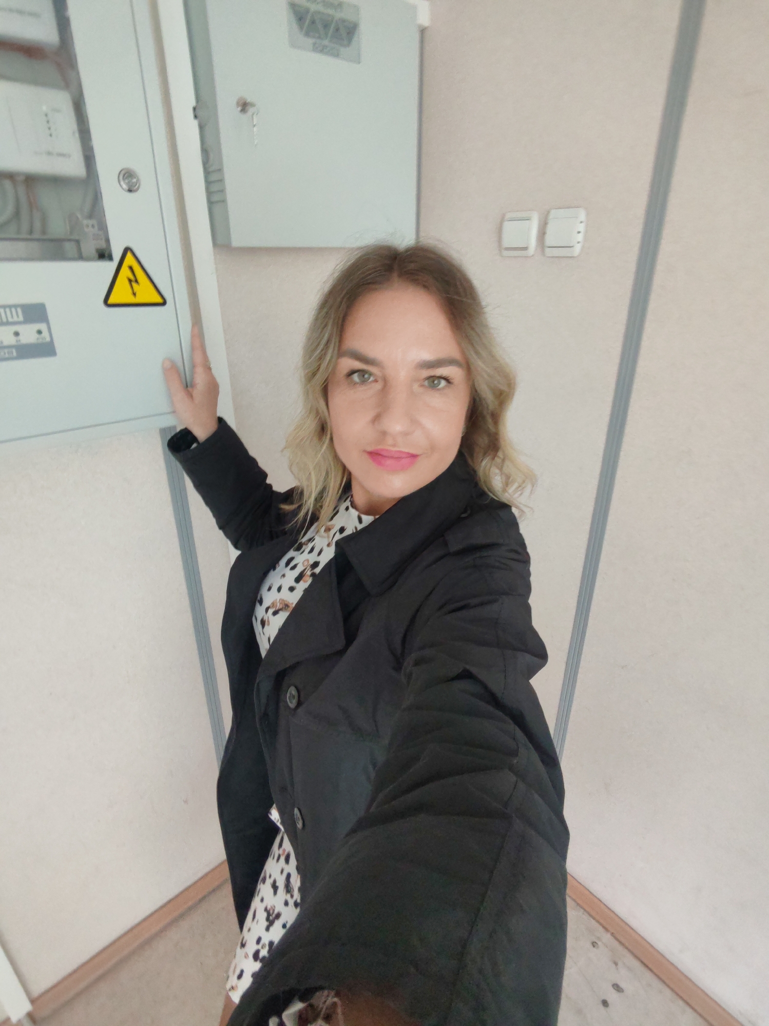 A woman stands in what appears to be an interior space, possibly a corridor or utility area, positioning herself next to an electrical panel. The panel is light gray and features a prominent yellow warning sign with a black lightning bolt symbol, indicating high voltage. The woman is wearing a black trench coat over a patterned dress. Her hair is blonde with dark roots. Two light switches are visible on the beige wall behind her. The location is unknown; the city is not evident in the image.