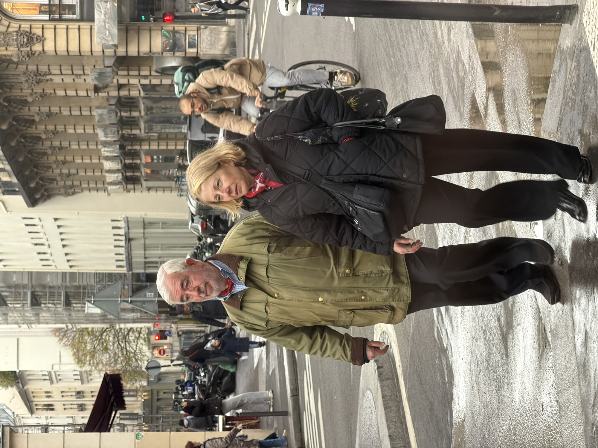 An eye-level outdoor medium shot presents an older Caucasian couple standing on a street in Paris, France. The man, on the left, has short white hair, light skin, and a slightly scruffy grey beard. He is wearing a light green jacket with brown cuffs over a light blue and white checkered shirt. A red scarf is visible at his neck. To his right, the woman has short blonde hair, light skin, and wears a black quilted jacket over a red and black patterned top. She carries a black shoulder bag. Both individuals are wearing dark pants and black shoes.
In the background, out-of-focus people are walking and riding bikes. Parked motorcycles and scooters line the street. The architectural style of the buildings in the background suggests Paris. Street signs and traffic lights are also visible. The wet street indicates recent rain. A portion of a black pole, possibly a street sign or lamp post, is visible in the upper right.