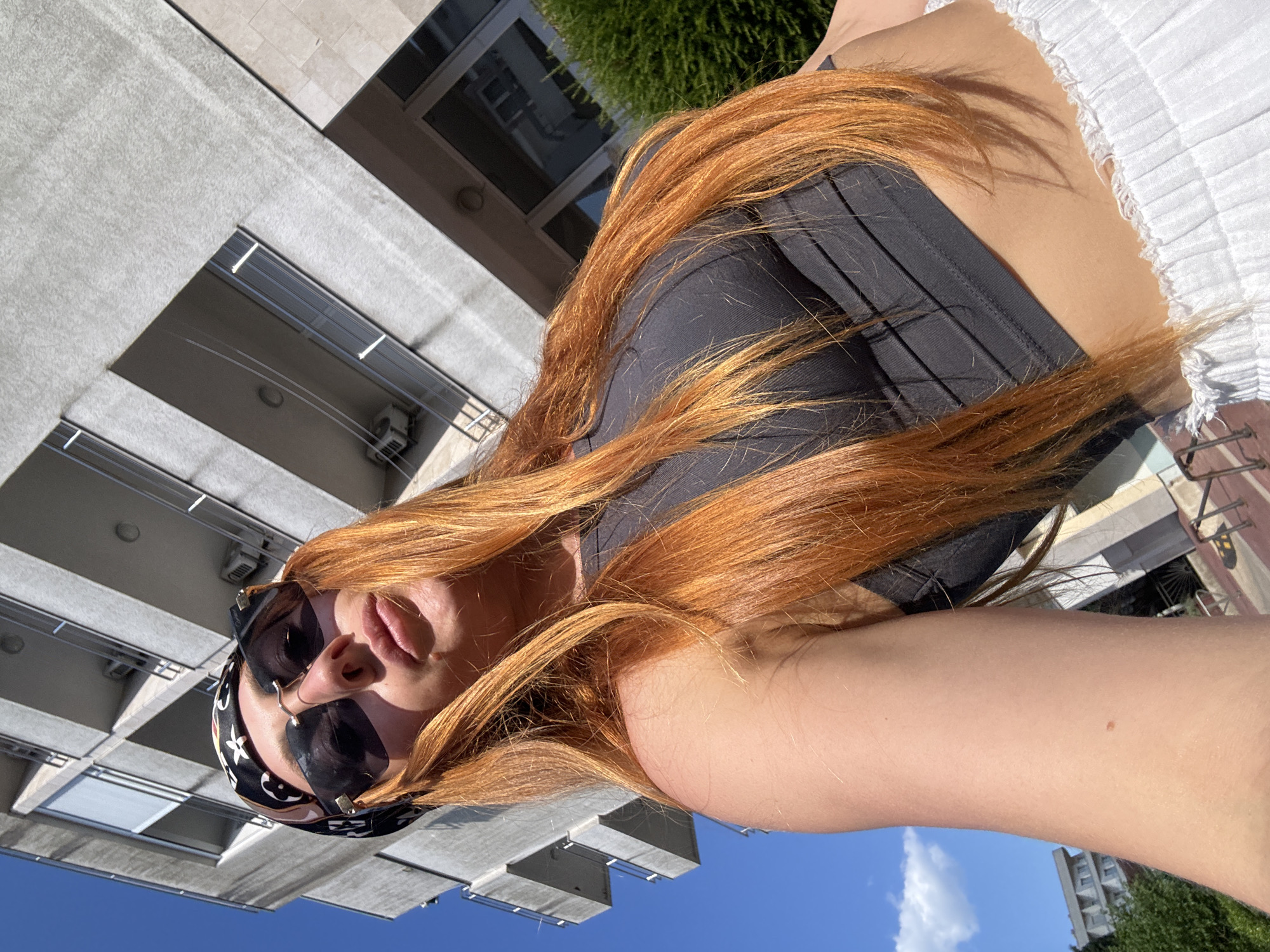 This is a rotated, close-up selfie of a person with long, auburn hair, wearing sunglasses and a black top. The person is outdoors, with a building and blue sky visible in the background. The building appears to be modern, with a concrete facade and balconies. Lush green foliage can be seen in the upper right corner. The bright sunlight suggests it is daytime, likely late afternoon or early evening given the warm, golden tones of the hair and shadows. The overall mood is casual and relaxed. No visible text is present in the image. The location is contextually identified as Budva, Montenegro.