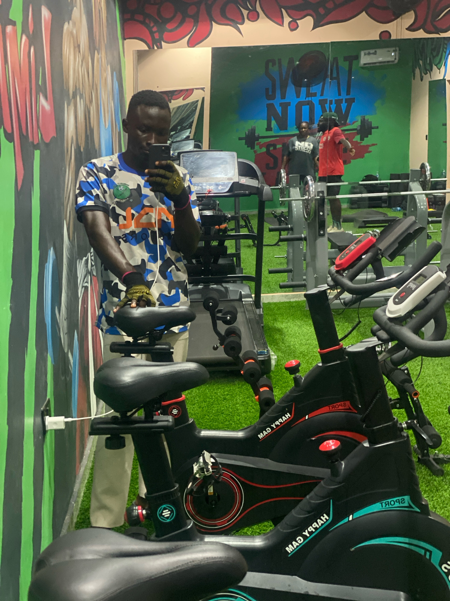 The image depicts the interior of a gym, likely located in Barden Yana, Nigeria. A young Black male is standing in the foreground, taking a mirror selfie. He is wearing a camouflage-patterned t-shirt, beige pants, and yellow workout gloves. He is holding a smartphone in his left hand and his right hand is resting on the seat of a stationary bicycle. Two stationary bicycles are visible in the foreground. In the background, through a mirror, other gym equipment such as treadmills and weightlifting racks are visible, along with two other individuals. The walls are decorated with graffiti art and motivational phrases like "SWEAT NOW." The floor is covered with green artificial turf. A white power adapter is plugged into an outlet on the wall, connected by a white cable to the base of the stationary bicycle in the foreground.