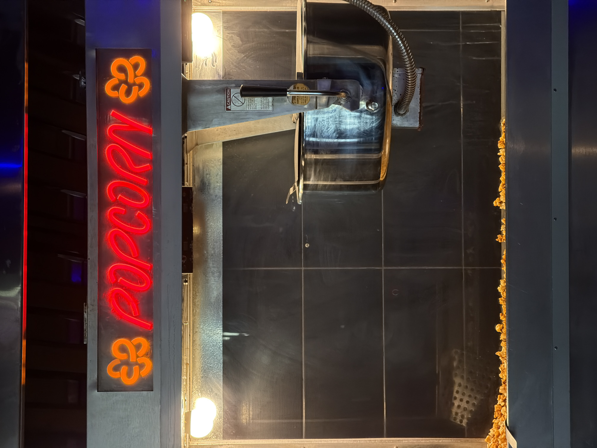 An indoor shot of a popcorn machine in Tbilisi, Georgia. The machine features a neon sign reading "POPCORN" vertically on its left side, flanked by what appear to be orange clover shapes. The machine itself is a large, metallic structure with a clear viewing window. Inside, one can see the popping mechanism above and a scattering of popcorn along the right edge, suggesting recent use. Two lights illuminate the interior. The environment is sleek and somewhat dimly lit.