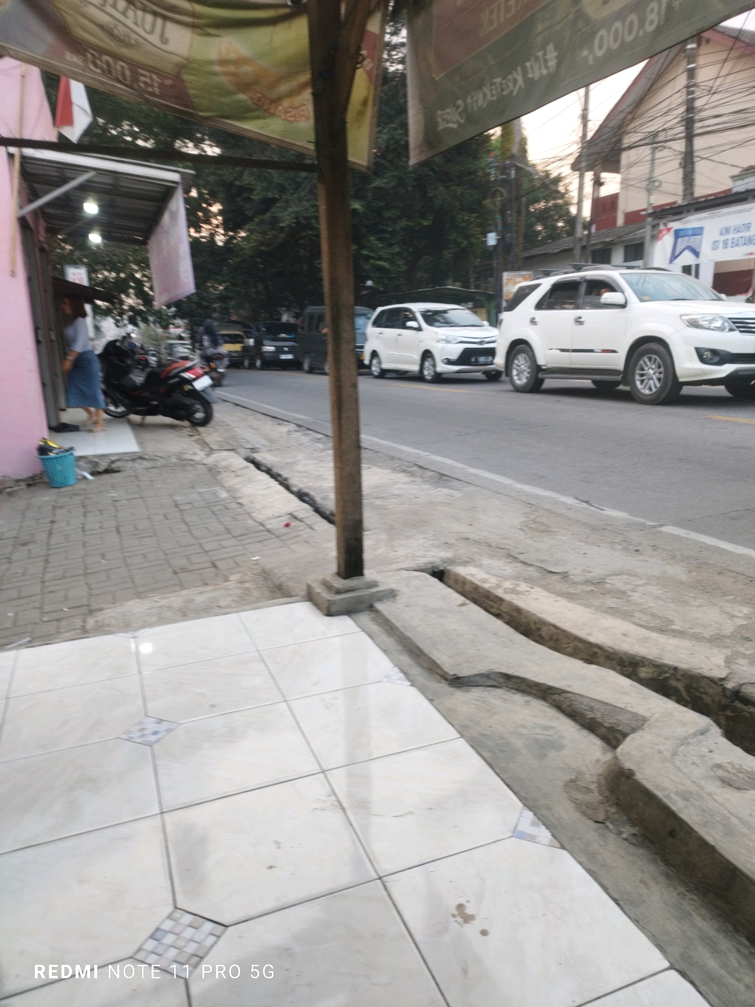 The image depicts a street scene in Saruni, Indonesia. In the foreground, a tiled walkway leads to a street with a curb and a drainage ditch. Beyond the ditch, a paved road is visible with several vehicles. A white SUV is parked on the right side of the road, and a white minivan is in motion on the left lane. Further down the street, other vehicles are present, including what appears to be a black van and motorcycles. On the left side of the image, there is a building with a pink facade, a canopy, and a blue trash bin. A person in a light-colored shirt and blue skirt is standing near the entrance of the building. Overhead banners are also visible, suggesting a commercial area. The overall environment appears to be a moderately busy street with shops and traffic. The sky is partly cloudy, and the lighting suggests it is daytime, possibly late afternoon or early evening.