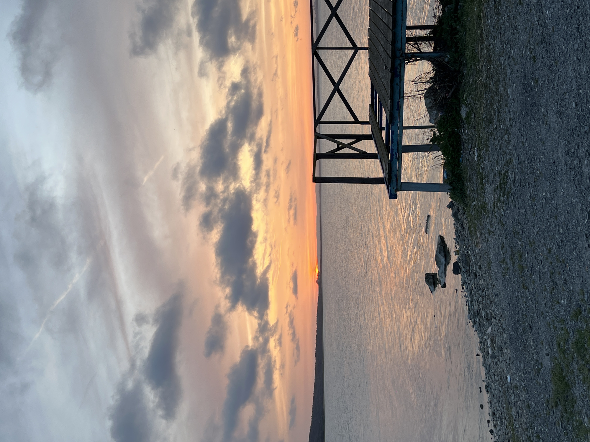 The image captures a serene sunset over a body of water, likely the Black Sea near Burgas, Bulgaria. The sky dominates the upper half of the frame, showcasing a beautiful blend of warm orange, pink, and pale blue hues, interspersed with soft grey clouds. The sun, a small, bright orb, is just visible above the distant horizon line on the left, casting a golden glow across the landscape. Several dark, scattered clouds add depth to the sky, contrasting with the illuminated areas.
The calm water below reflects the vibrant sky, shimmering with golden light from the setting sun. On the right side, a sturdy wooden structure, possibly a pier, dock, or a large bench, extends partially over the water. Its dark silhouette, featuring crisscrossing support beams, stands out against the reflective surface.
The immediate foreground on the right is composed of a gravelly or rocky shore, with sparse patches of green grass and some visible debris or small rocks. A few larger rocks are partially submerged in the water close to the shoreline. The overall scene evokes a sense of tranquility and peacefulness as day transitions to night. There is no visible text in the image.