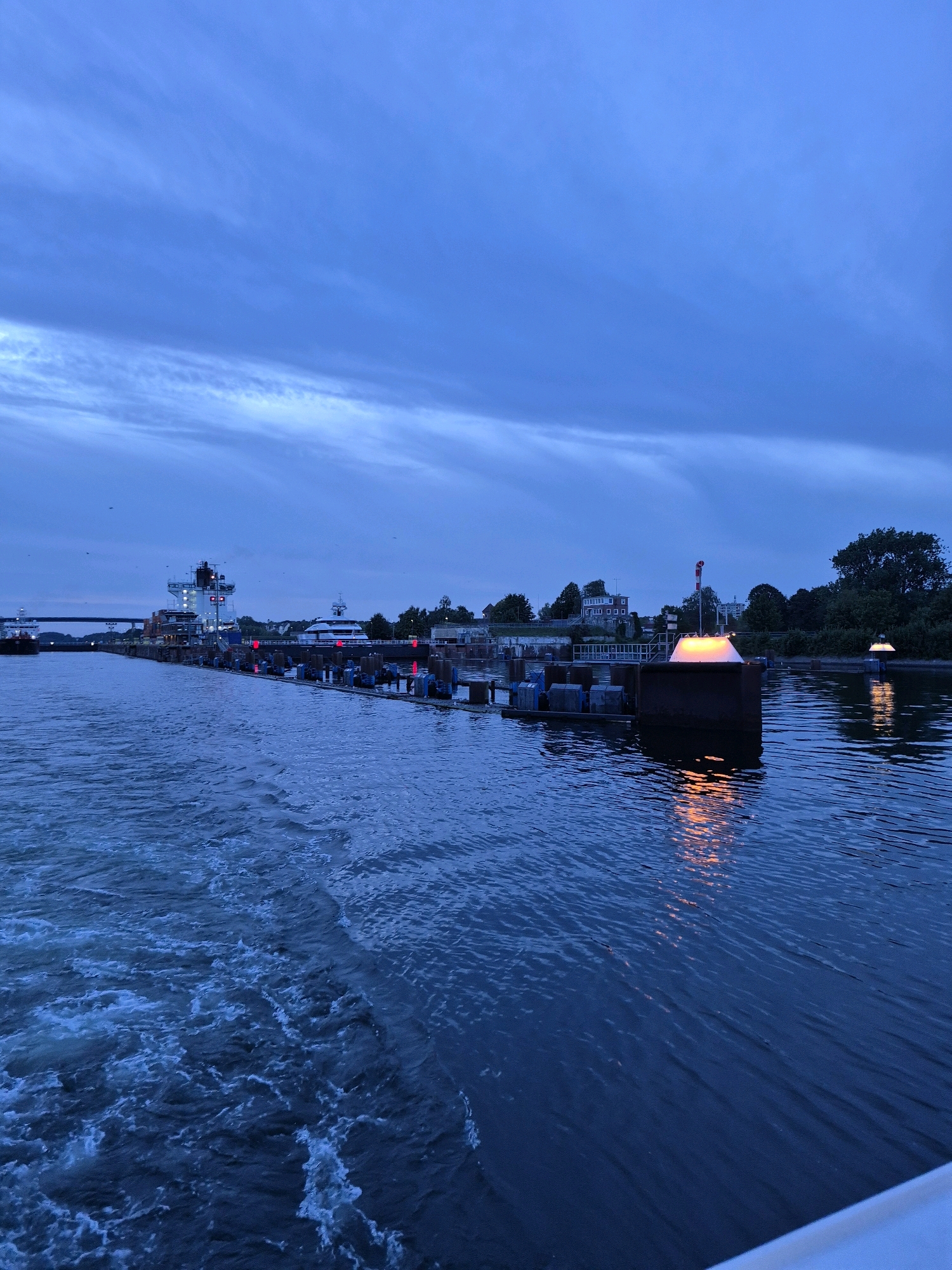 The image depicts a wide waterway, likely a canal or river, at dusk or dawn. The water is a deep blue, with some churning white wake on the left side of the frame, suggesting movement. Along the right bank, there is a constructed embankment with a lock system. Several barges or pontoons are docked, some of which have bright, orange lights illuminating them and reflecting on the water. Beyond the embankment, trees and buildings are visible, including a large, modern white boat docked on the right. Further down the waterway on the left, a large cargo ship is docked, its superstructure illuminated with red and white lights. A bridge spans the waterway in the distance. The sky above is a deep blue, with streaks of lighter clouds. The scene appears to be a port or industrial area. Given the context of Kiel, Germany, this scene could be the Kiel Canal or the harbor area.