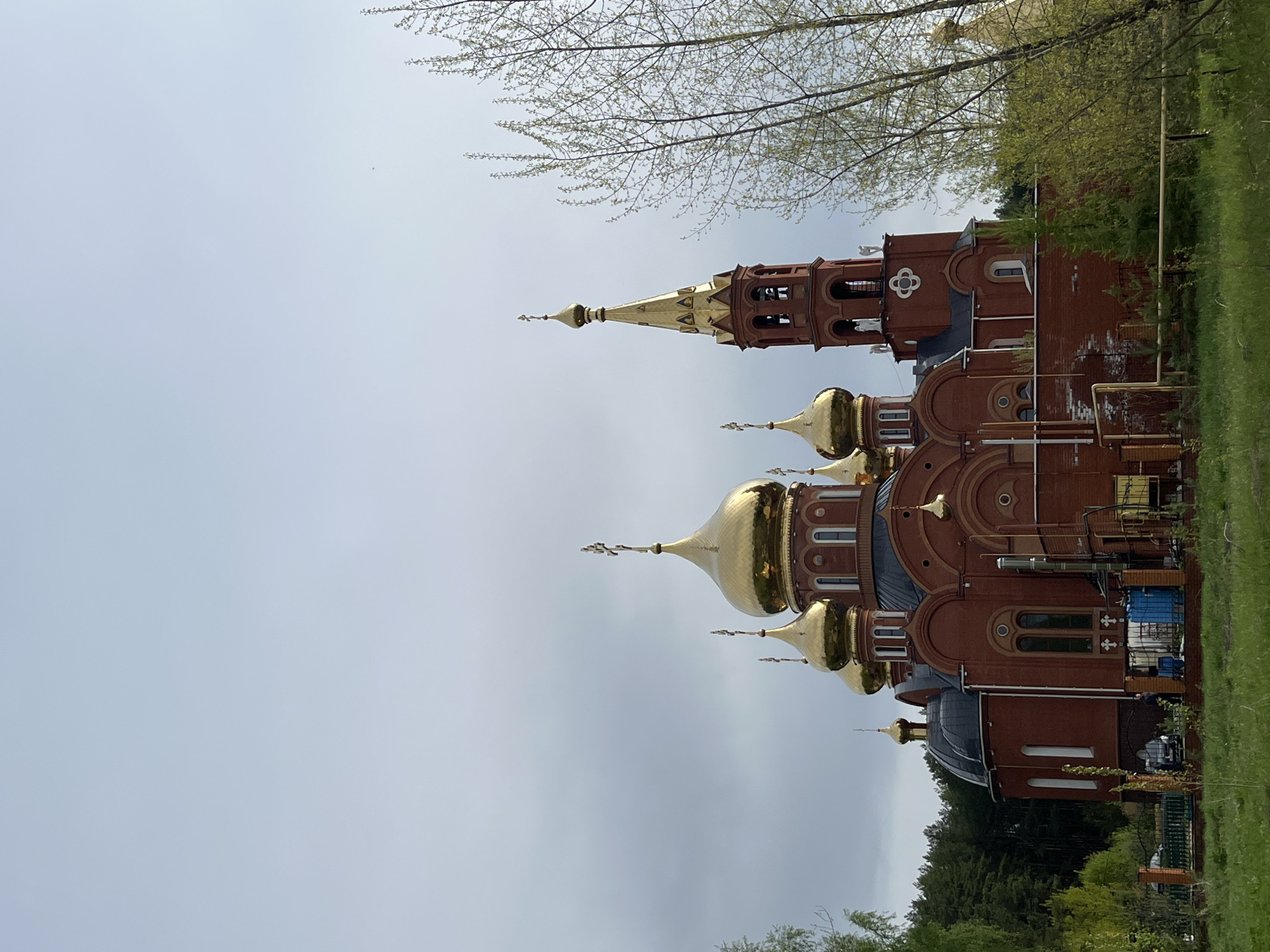 The image shows the Church of the Holy New Martyrs and Confessors of Russia, located in Chaikovsky, Russia. The church is a red brick building with multiple golden domes and crosses. A bell tower is visible. The sky is overcast, and trees are present in the foreground.