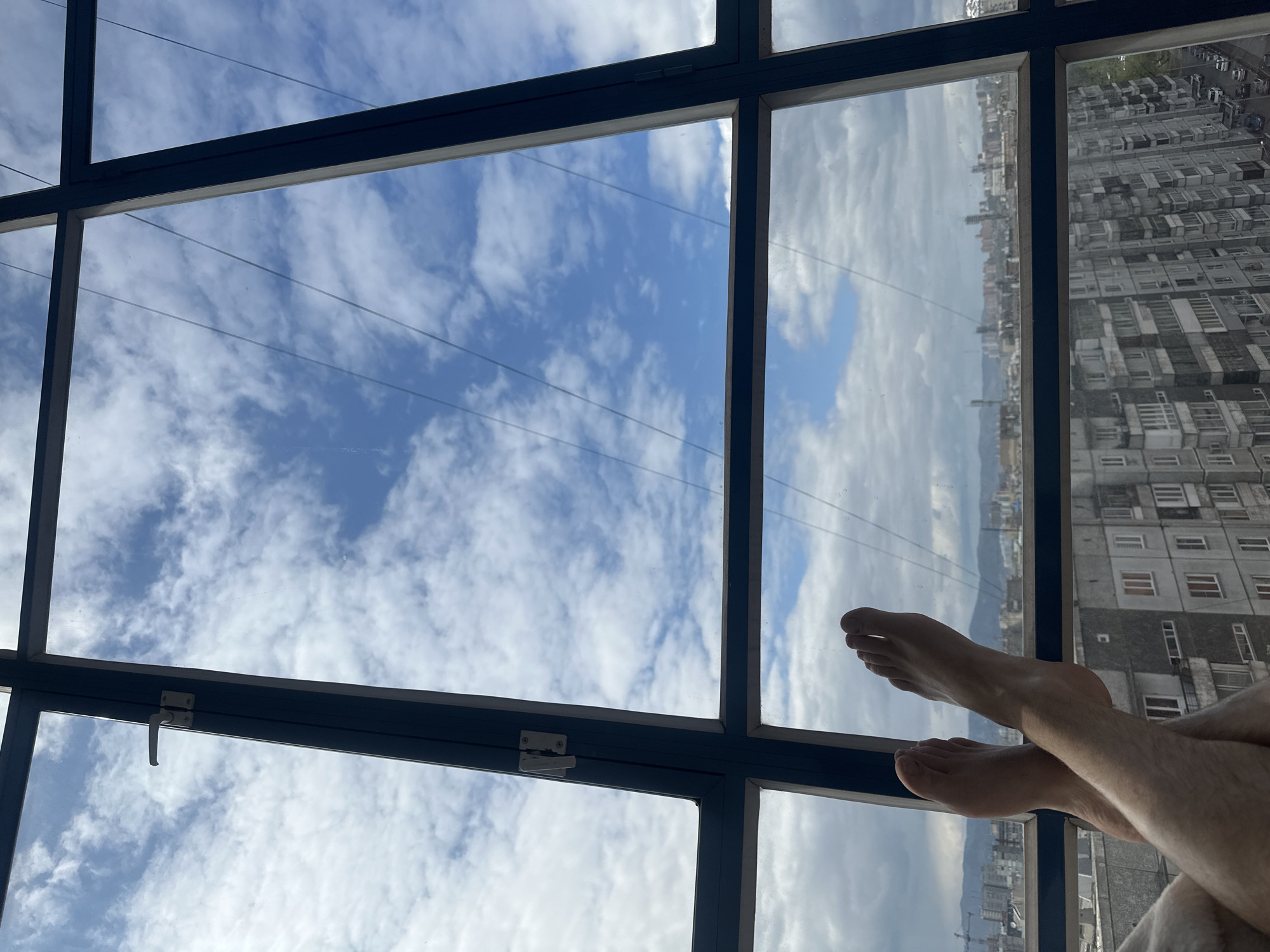 The image shows a view from inside a room, looking out through a multi-paned window. The sky is visible through the window, with scattered clouds against a blue background. In the distance, a cityscape is visible. The legs and feet of a person are also visible, suggesting someone is relaxing and looking out the window.
The location is Krasnoyarsk, Russia. The buildings in the background appear to be typical apartment blocks.