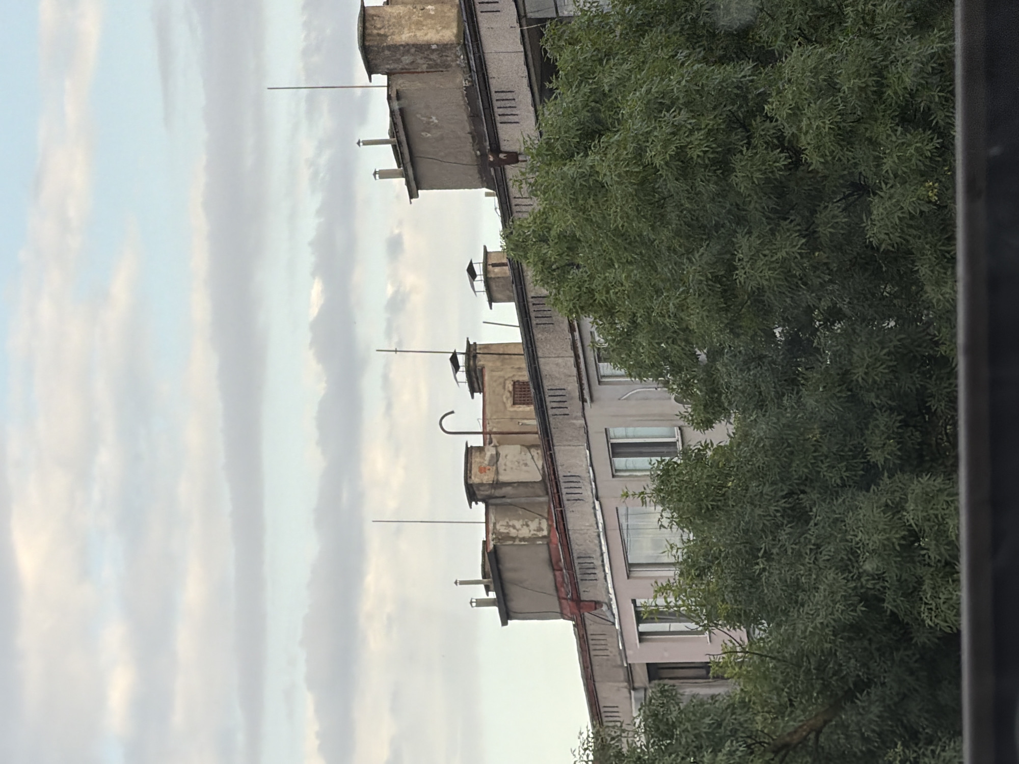 This image captures an urban scene in Ruse, Bulgaria, featuring the upper floors of residential buildings and a cloudy sky. The buildings are utilitarian in design, with visible concrete structures, chimneys, and ventilation pipes on their flat roofs. Some buildings show signs of wear, with peeling paint and discolored facades. The architecture suggests an older construction style common in Eastern European cities. Lush green trees with dense foliage partially obscure the lower levels of the buildings, indicating a residential area with established greenery. The sky is overcast with diffused light, creating a muted atmosphere. There are no people visible in the frame. The perspective is from a slightly elevated viewpoint, looking across a street or open space towards the buildings.