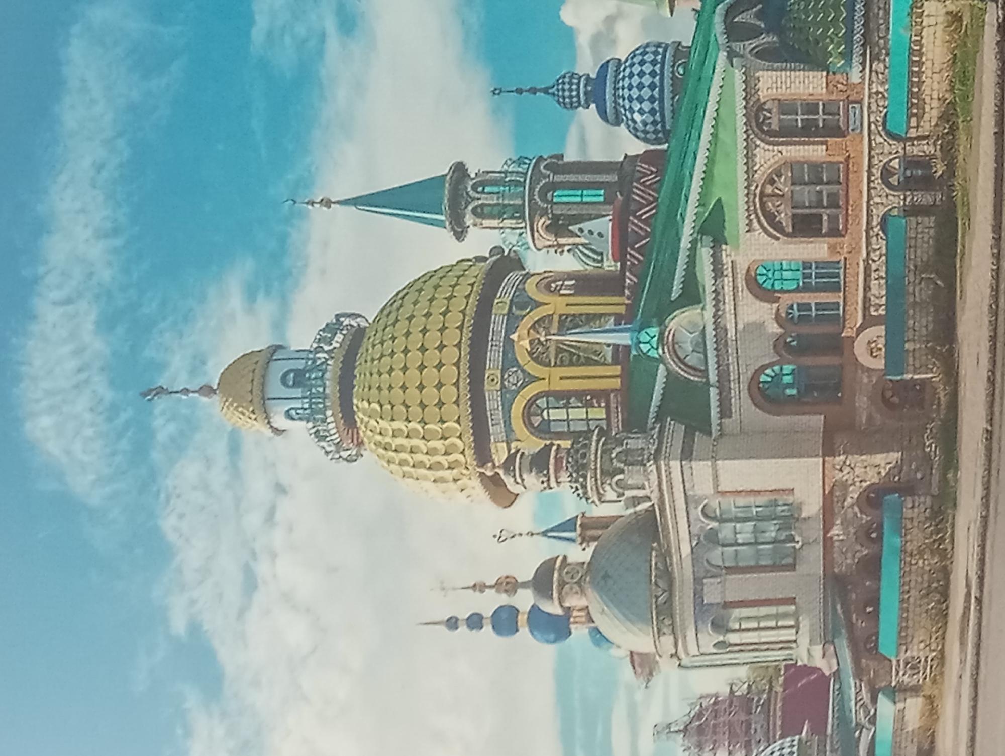 The image shows a colorful, ornate building complex under a partly cloudy sky. The architecture includes multiple domes of different sizes and colors, with one prominent gold-colored dome. There are also towers, spires, and arched windows visible. The building has a variety of architectural styles mixed together. The image is of the Temple of All Religions, located in Karaganda, Kazakhstan.