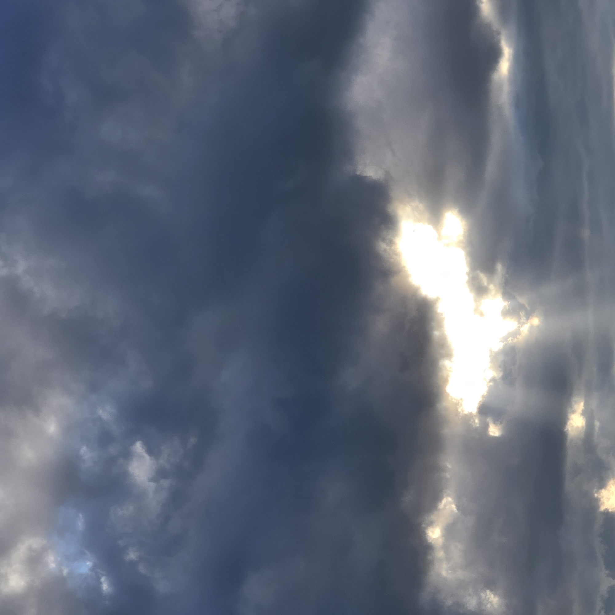 The image shows a dramatic sky filled with dark, ominous clouds. A bright, irregular patch of light breaks through the clouds in the lower-middle portion of the frame, with visible rays of sunlight emanating downwards. The clouds are textured and appear dense, with varying shades of gray and deep blue, suggesting stormy or overcast conditions. The sunlight piercing through creates a stark contrast, illuminating parts of the cloud edges and casting a soft glow. The scene is devoid of any man-made structures or identifiable geographical features of Belokurikha, Russia, and contains no people or animals. It is a purely atmospheric depiction of a sky with intense light and shadow play.