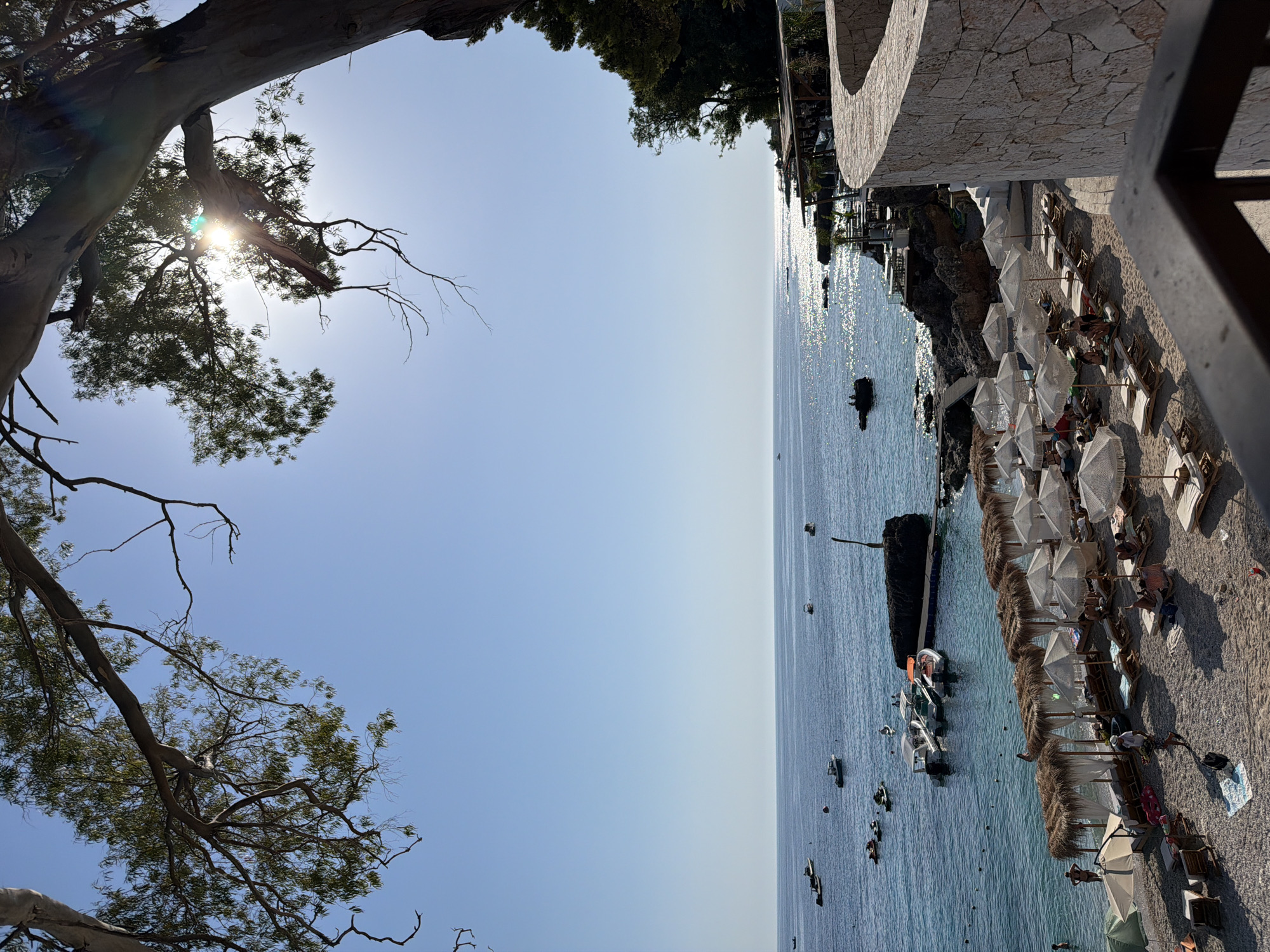 The image shows a sun-drenched beach scene in Himarë, Albania. A calm, blue sea stretches to the horizon, dotted with several small boats and jet skis. The shoreline is a mix of sand and pebbles, with a section dedicated to beach chairs and umbrellas. Many of these are arranged under thatched-roof cabanas, offering shade to beachgoers. Several people are visible lounging on the chairs or walking on the beach. In the background, rocky outcrops jut out into the sea, and lush green trees line the coast. A curved stone wall borders the right side of the image, suggesting a pathway or elevated viewing area. The sun is bright, casting a shimmering reflection on the water, and large tree branches with green leaves frame the top of the scene, partially obscuring the clear blue sky.