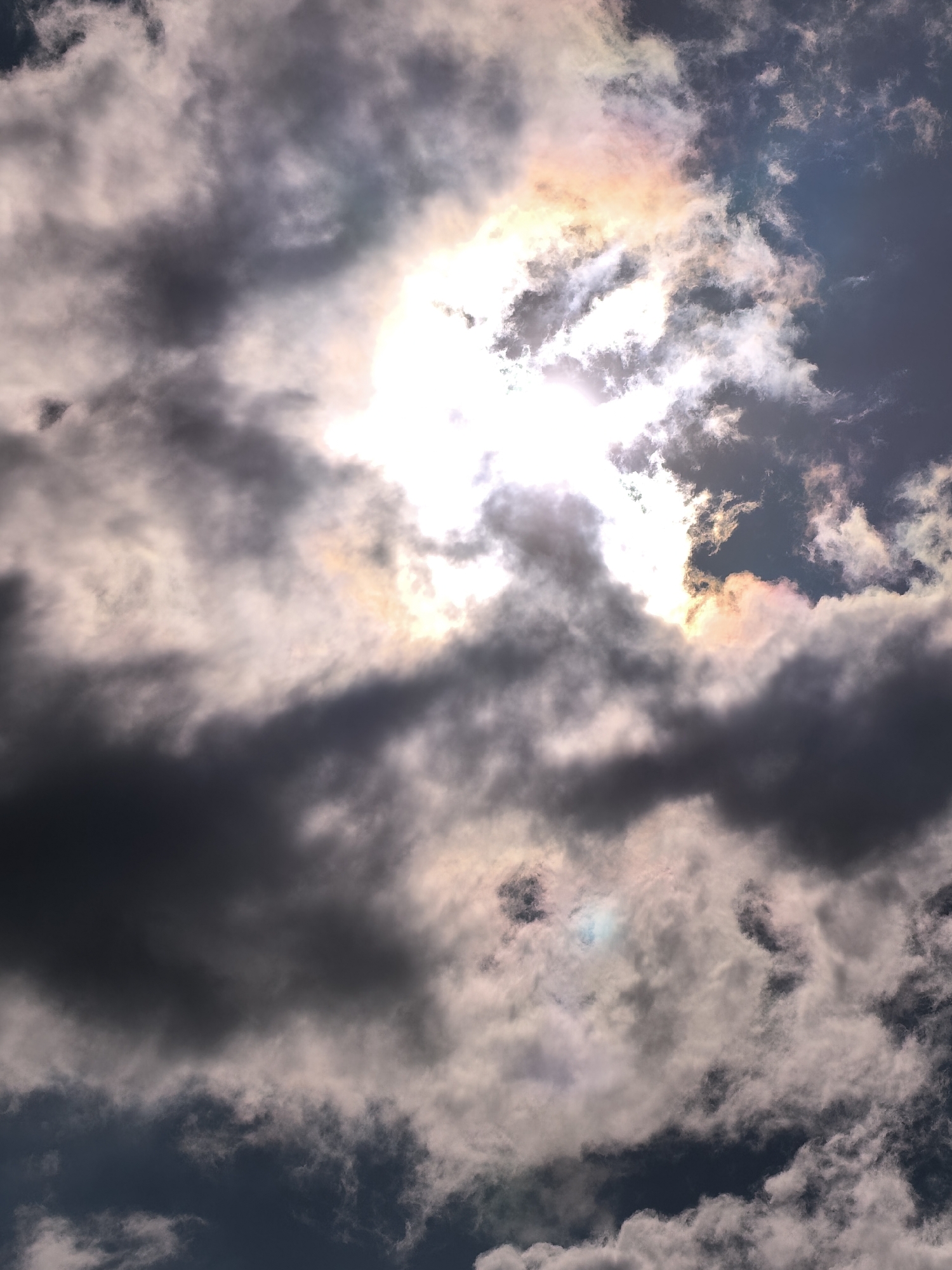 The image shows a sky filled with dark, ominous clouds. The sun is partially obscured by the clouds, creating a bright, diffused light with a hint of rainbow iridescence around the edges. The scene is located in an unknown city. There are no people or other objects visible in the image.