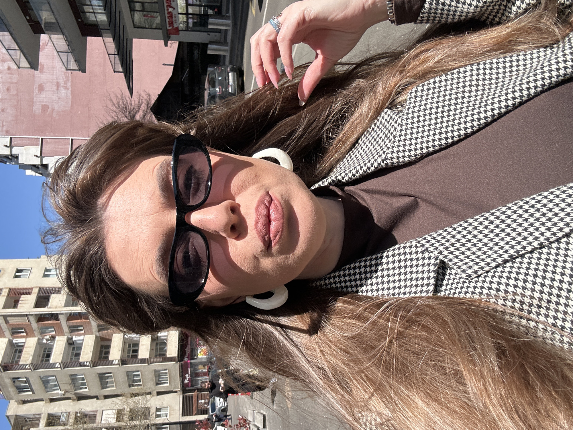 The image shows a woman with long brown hair, wearing sunglasses and large white hoop earrings. She is also wearing a brown top and a black and white houndstooth blazer. Her right hand is raised near her head. Buildings are visible in the background. The location is Tbilisi, Georgia.