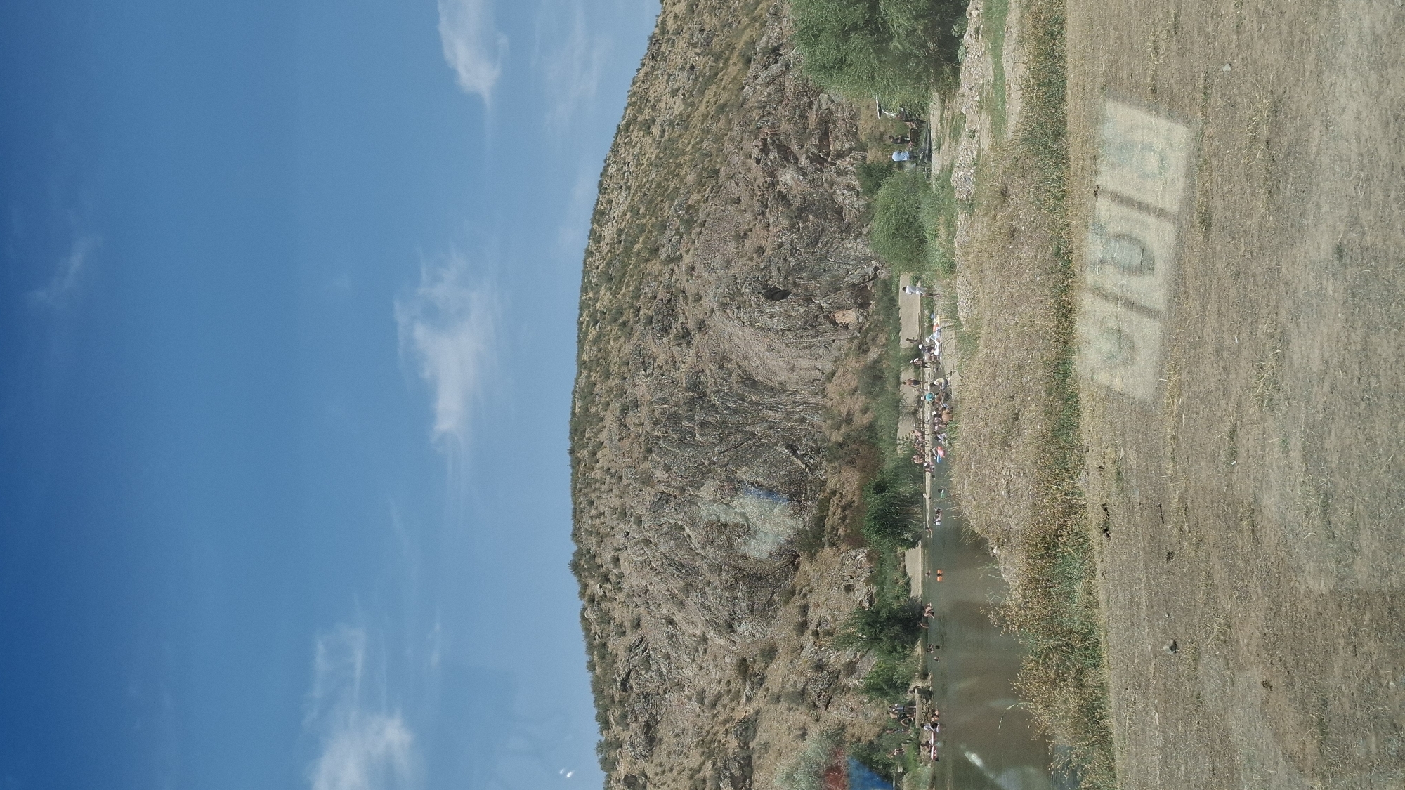 This image depicts a scene at a body of water, likely a river or lake, on a sunny day. In the foreground, dry, dusty ground with sparse vegetation is visible. The middle ground features the body of water, with numerous people scattered along its banks and within the water. Many individuals are swimming, wading, or relaxing on the shore, some appearing to be on inflatable devices. Lush green vegetation, including reeds and trees, lines the water's edge.
Dominating the background is a large, rocky, arid hill with sparse bushes and vegetation. The hill's surface is textured and appears to be composed of layered rock formations. The sky above is a clear blue with a few scattered white clouds. A section of the rocky hill exhibits a patch of blue and light grey coloring, possibly due to mineral deposits or a colorful rock formation. The overall environment suggests a natural, recreational area.
Based on information about the region, this scene is likely located at the Boralday resort area near the city of Boralday, Kazakhstan, known for its natural landscapes and recreational opportunities.