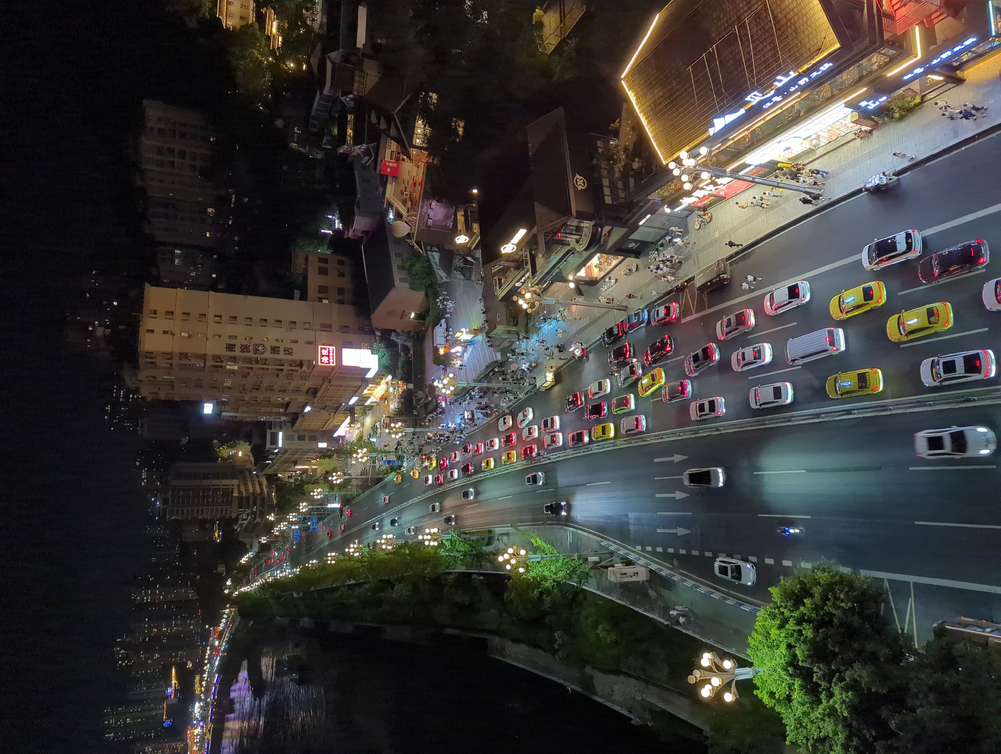 The image captures a nighttime view of a busy multi-lane road alongside a river in Yuzhong District, China. Numerous cars, predominantly in red, yellow, and white, are traveling in both directions, their headlights and taillights creating streaks of light. Pedestrians are visible on the sidewalks lining the road, some clustered in groups, others walking individually. Buildings of varying heights, illuminated by interior lights and exterior signage, form the urban backdrop, with some structures exhibiting traditional architectural elements. Streetlights with multiple globes line the roadside, casting pools of light on the street and sidewalks. The river, on the left side of the frame, reflects the lights from the city, creating a shimmering effect. The overall scene depicts a vibrant urban environment at night, characterized by continuous movement and illumination.