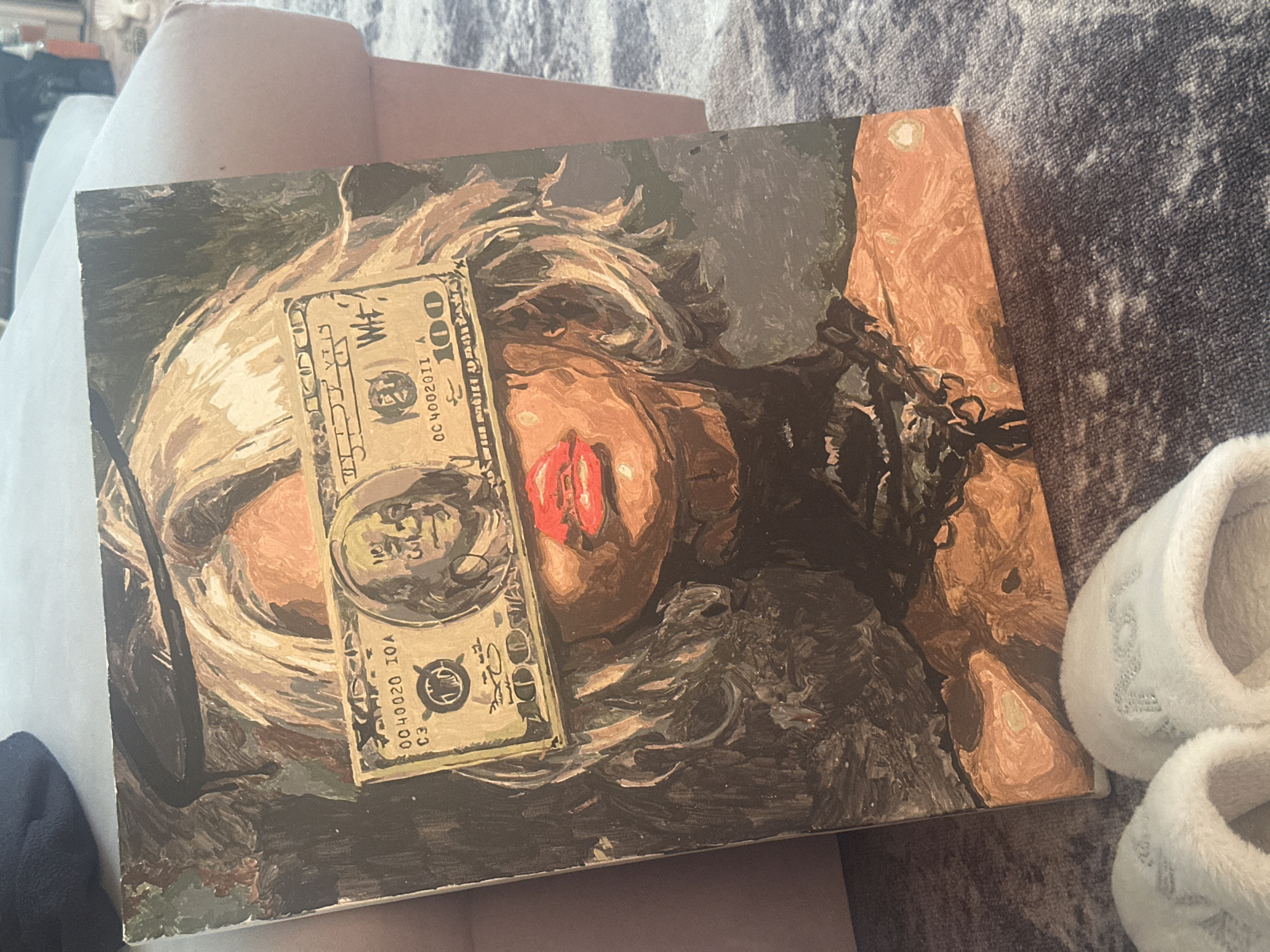 The image shows a painting resting on a soft surface (likely a cushion or sofa). The painting depicts a woman with blonde hair partially obscuring her face, red lipstick, and what appears to be a dark, undefined mass around her neck and shoulders. A US $100 bill is superimposed over the woman's face, partially covering her right eye and cheek.
There is no information in the image to associate it with the city of Высокий, Russia.