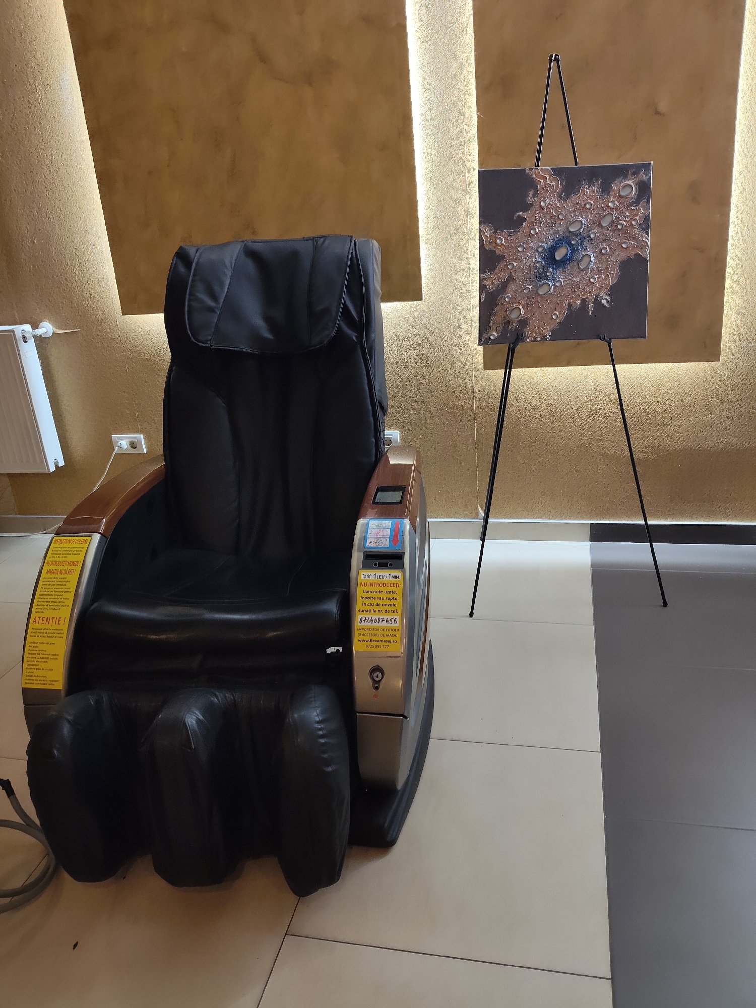 An indoor scene depicts a black massage chair positioned to the left of a painting displayed on an easel. The painting, square in shape, features an abstract design with blues, browns, and metallic elements. Behind the massage chair and the easel, a textured, golden-brown wall is illuminated by indirect lighting from above. To the far left, a white radiator is visible against the wall. The floor is tiled with large square tiles, with a transition to darker grey tiles on the right side of the frame. The massage chair itself has wooden armrests and a control panel on the right side, adorned with stickers in Romanian. The lower portion of the chair features inflated leg rests.
