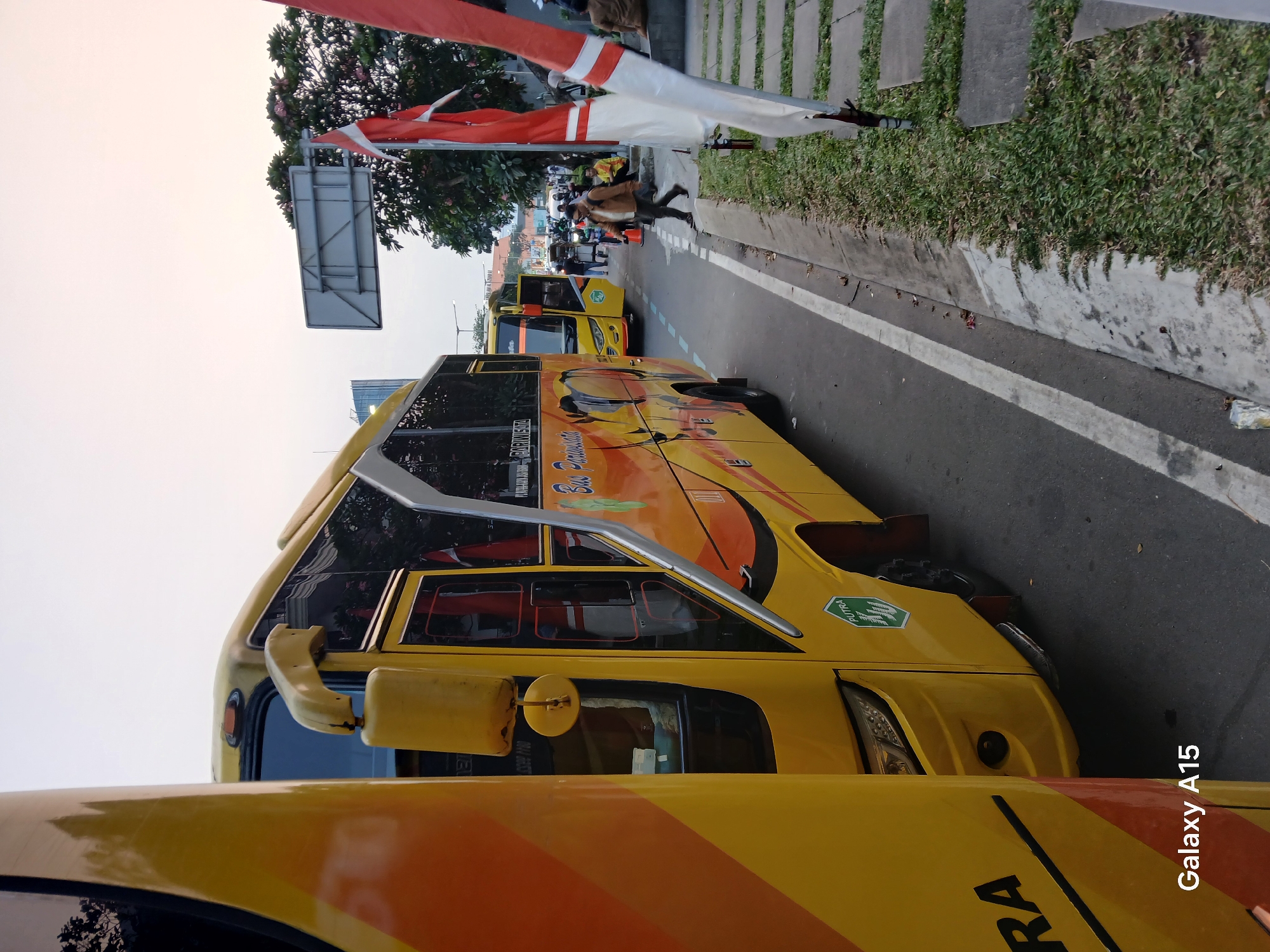 In Curug Sangereng, Indonesia, a yellow tour bus with "Bus Pariwisata" branding is parked on the side of a street. The bus has a distinctive yellow and orange design with a graphic of a bird on its side. In the foreground, another yellow bus is partially visible on the left. Several people are present near the bus, with some walking and others sitting on a stone bench. Indonesian flags are displayed on poles in the foreground. The sky appears overcast, suggesting it might be early morning or late afternoon. A signpost is visible in the background. The scene depicts a typical street view with vehicles and pedestrians.