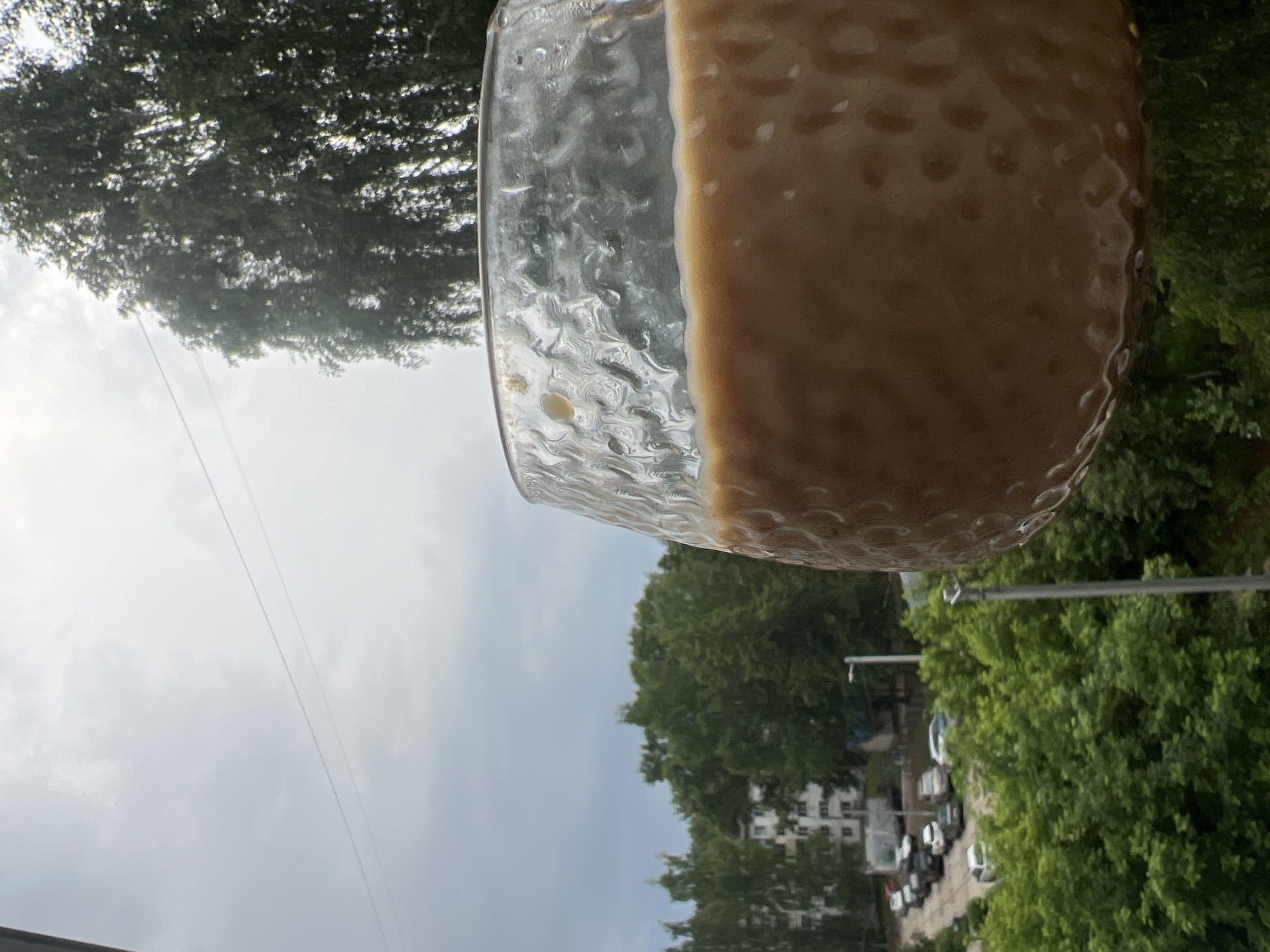 A close-up, low-angle shot captures a glass of light brown liquid, possibly coffee or tea with milk, set against a hazy, overcast sky. The glass has a textured surface with a dimpled pattern and condensation droplets. In the background, a line of parked cars is visible on a street lined with lush green trees. Beyond the trees, a multi-story residential building with white facades stands, indicating an urban or suburban setting. The overall scene appears to be in Chaikovsky, Russia, given the architectural style and vegetation.