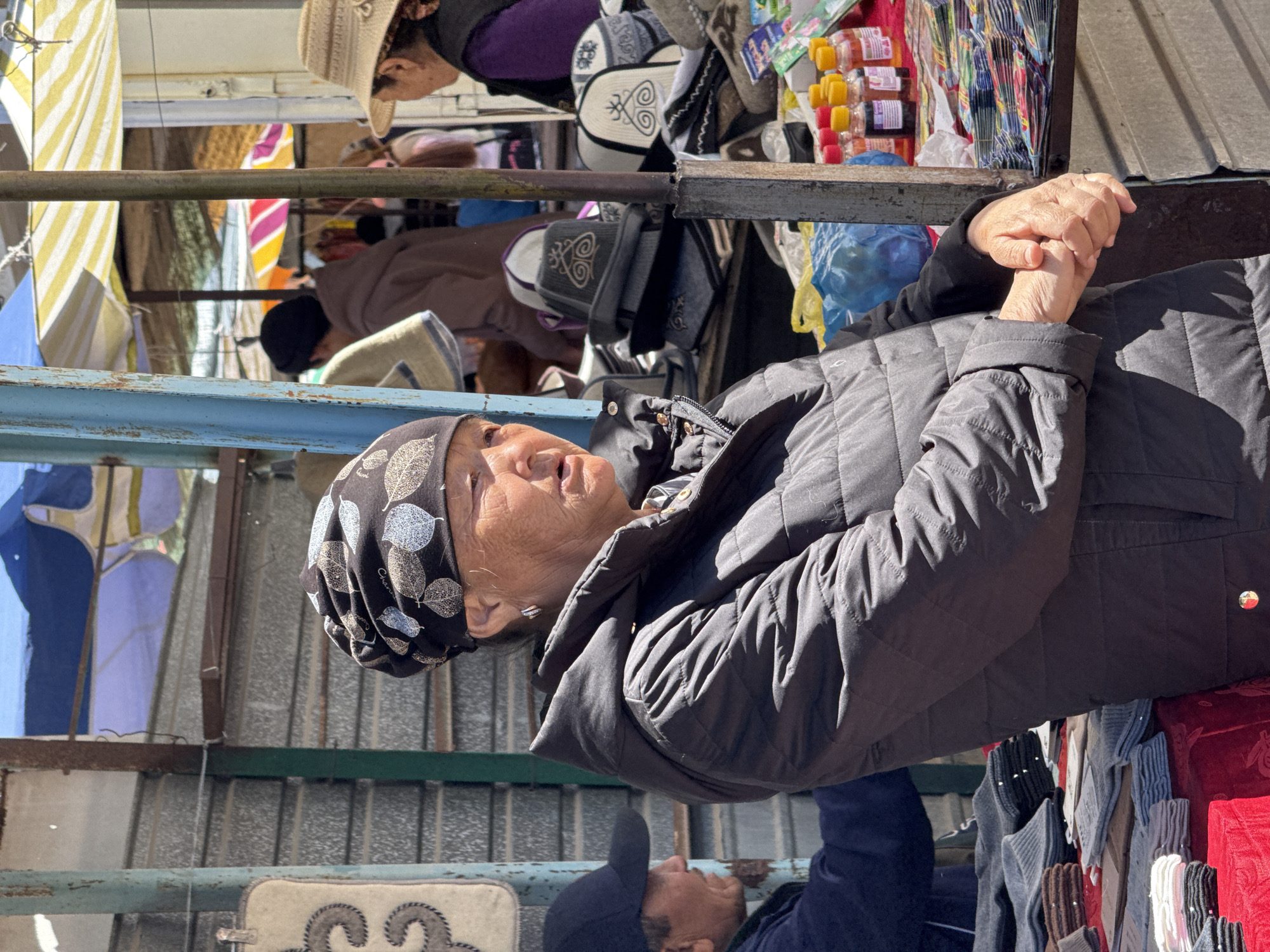 An elderly woman wearing a black headscarf with a leaf pattern and a black quilted jacket stands in a marketplace. Her hands are clasped together, and she is looking to her left. Behind her is a metal support structure with various market stalls and items for sale, including hats, clothing, and bottled goods. Another person wearing a blue baseball cap is visible in the foreground. The location is likely in a city market in Bosteri, Kyrgyzstan.